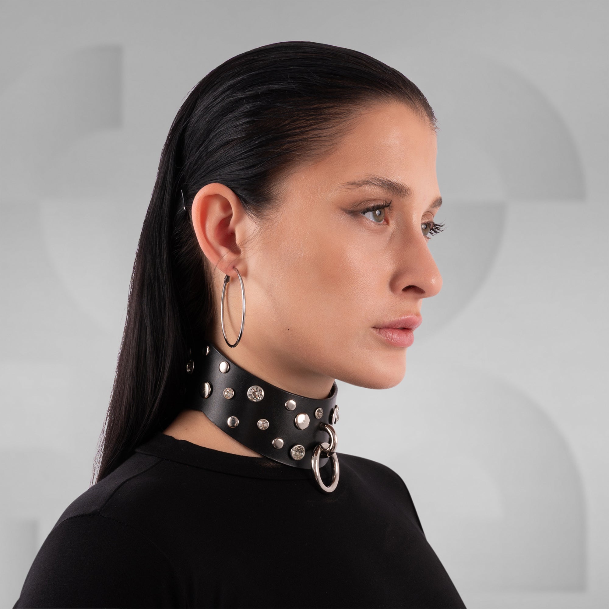 Girl wearing a black leather choker with glas stones and siver rings in front of a grey background
