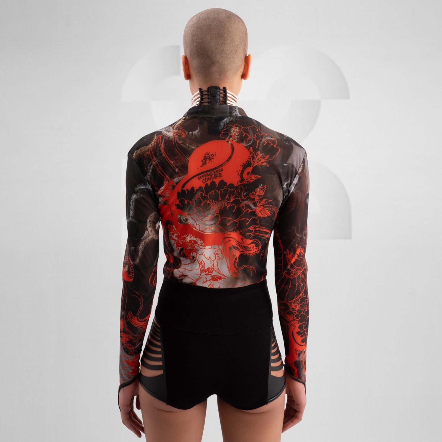 The image showcases a striking outfit featuring a long-sleeve top with an eye-catching design of swirling octopuses in dark hues, accented with vibrant red. The top is complemented by unique metallic neck detailing that adds an edgy flair. Paired with black high-waisted shorts featuring cut-out details, this look exudes confidence and individuality. Ideal for making a statement on the dancefloor or at a fashion-forward event, the ensemble beautifully blends art and style for a captivating appearance.