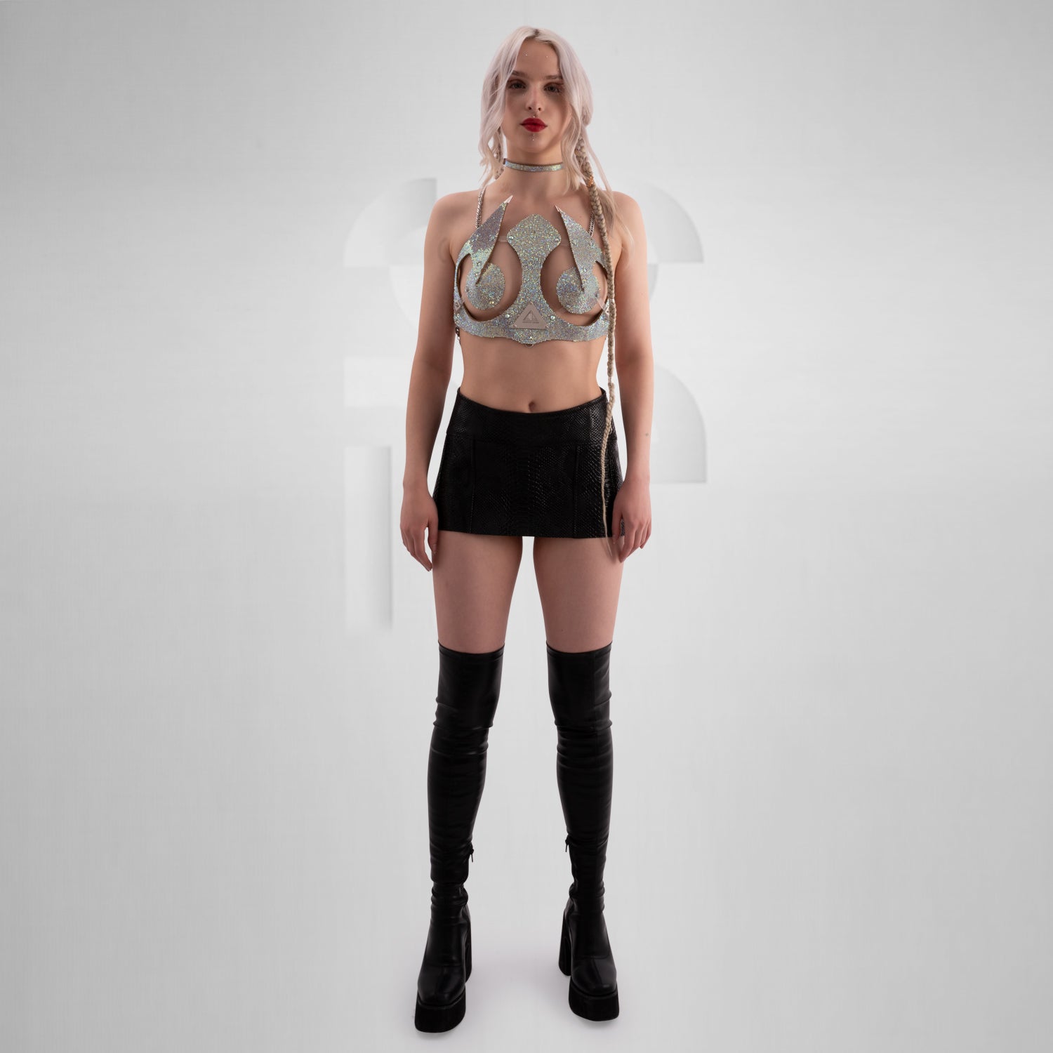 This image features a woman with platinum blonde hair styled in loose waves and a single braid on one side. She is wearing a highly decorative and futuristic top made of shimmering, rhinestone-encrusted materials that cover her chest and shoulders, with pointed embellishments extending outward. Her makeup includes bold red lipstick, and she has multiple facial piercings, including a septum ring and dermal piercings near her eyes and lips.