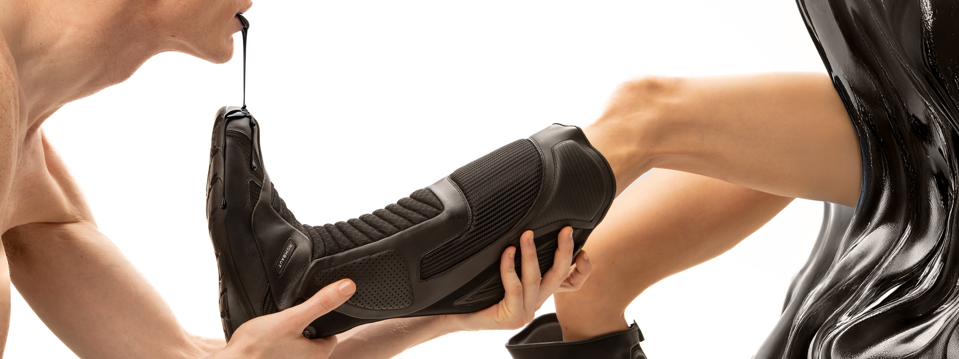 Scene featuring two models interacting, one holding a black shoe while the other extends their leg. Black liquid is shown dripping, creating a striking visual effect. The focus on the footwear and the unique textures enhances the edgy aesthetic of the composition, set against a clean white background that emphasizes the details and contrasts.