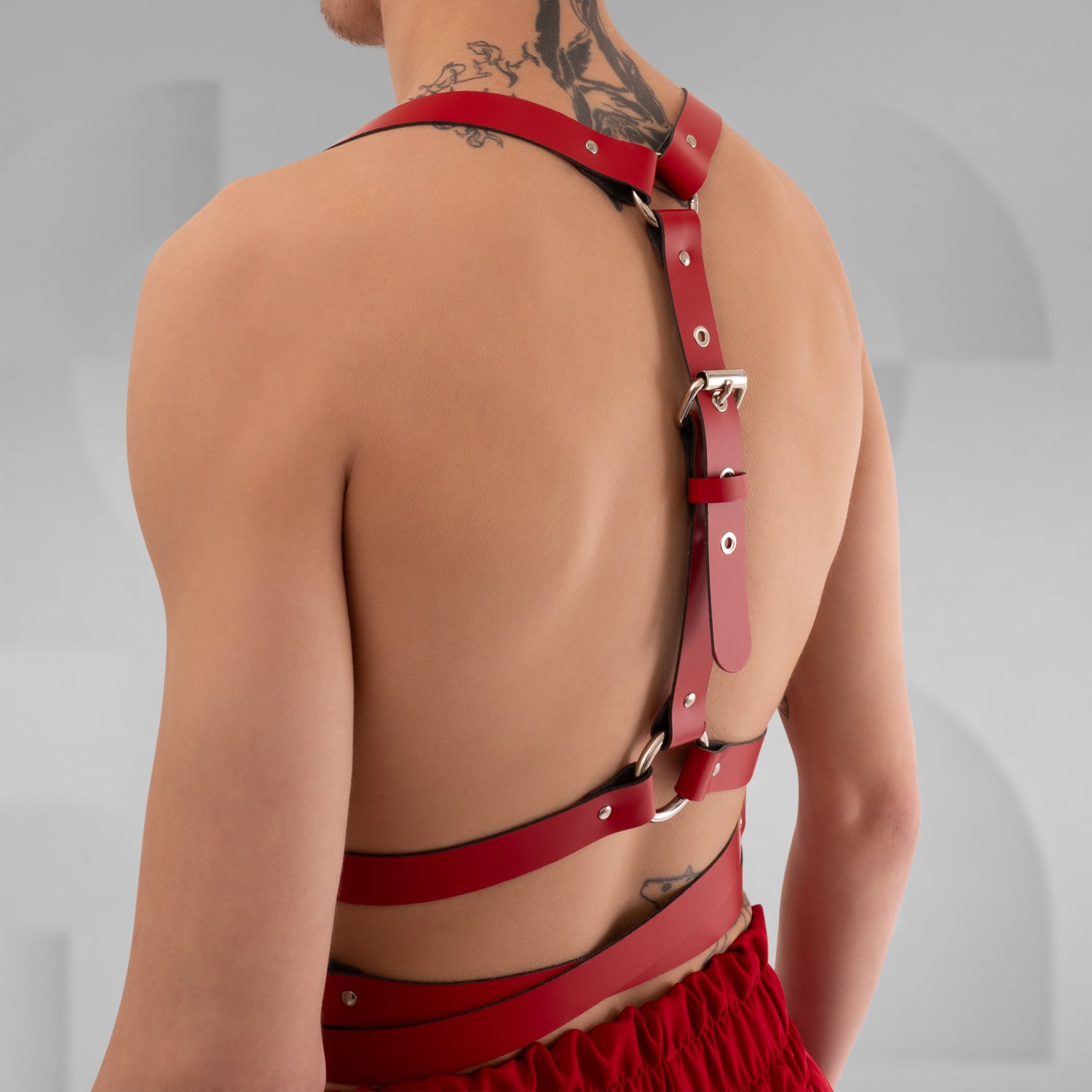 a stylish red leather harness with crisscross straps and metal hardware, creating a bold and modern look. The minimalist design is complemented by the neutral background, highlighting the harness's unique features and the model's confident stance.