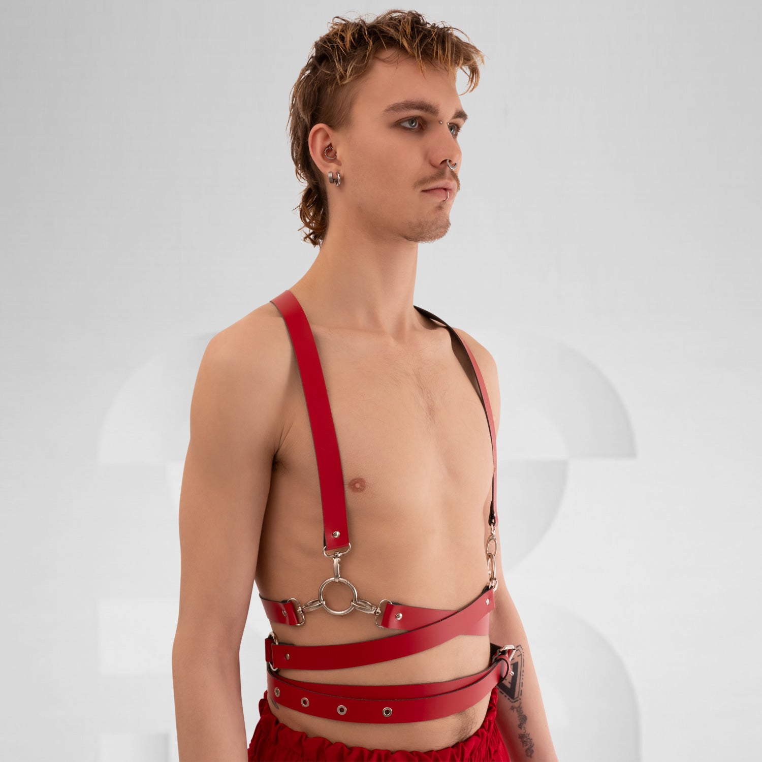 a stylish red leather harness with crisscross straps and metal hardware, creating a bold and modern look. The minimalist design is complemented by the neutral background, highlighting the harness's unique features and the model's confident stance.