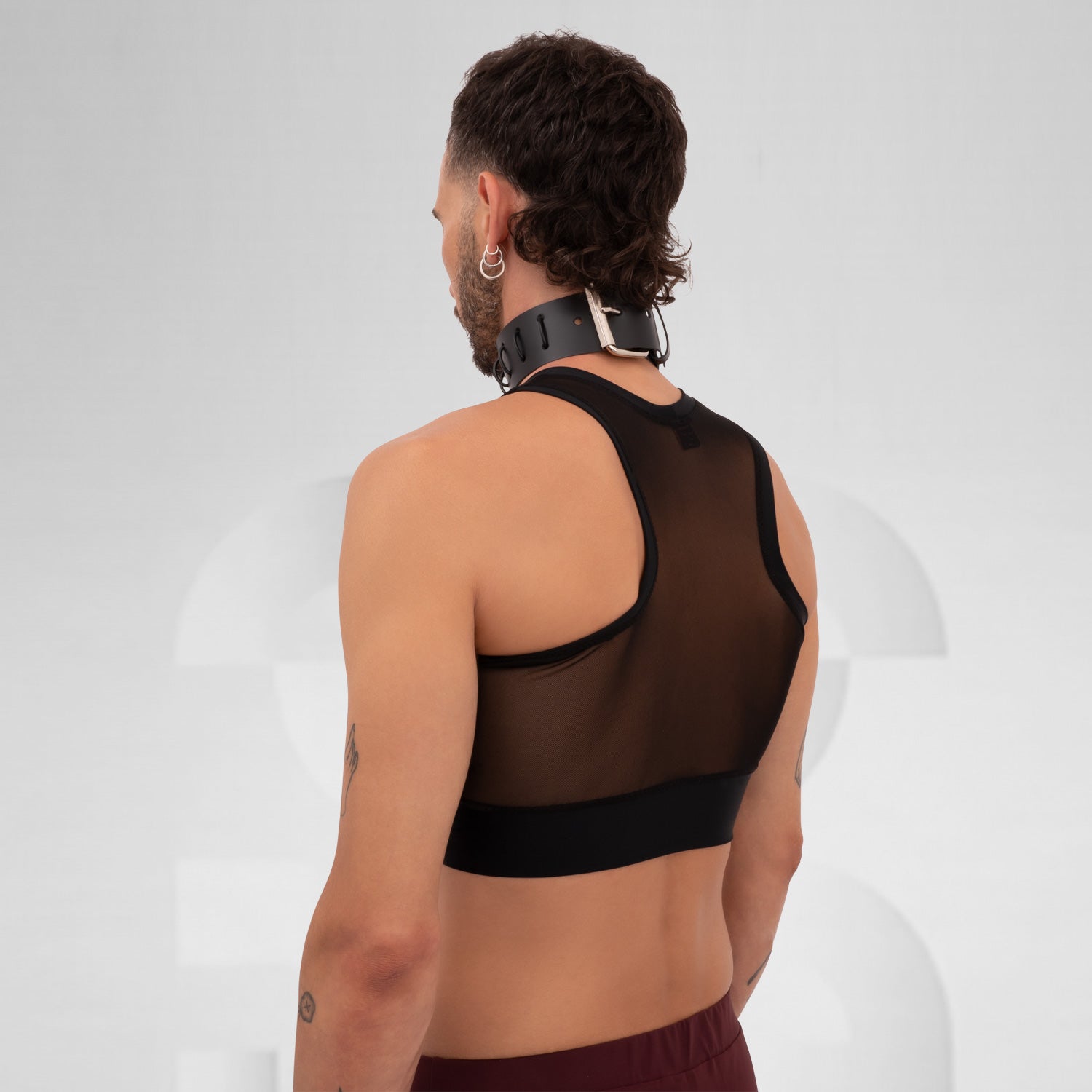 The a model wearing a black, sheer top with a sporty cut, featuring a layer of transparency. The outfit is accented by a chunky choker with a metallic detail. The model's arms are tattooed, showcasing various designs that add character to the look. The neutral background draws focus to the ensemble and the model's confident expression.