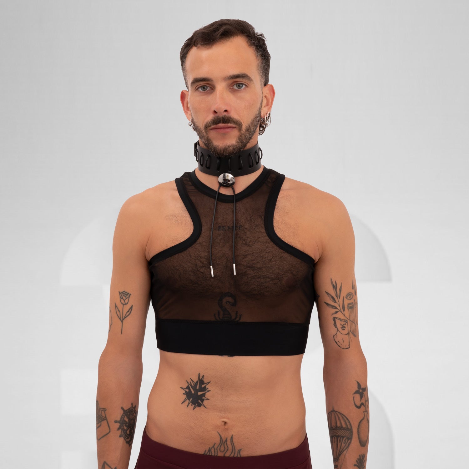 The a model wearing a black, sheer top with a sporty cut, featuring a layer of transparency. The outfit is accented by a chunky choker with a metallic detail. The model's arms are tattooed, showcasing various designs that add character to the look. The neutral background draws focus to the ensemble and the model's confident expression.