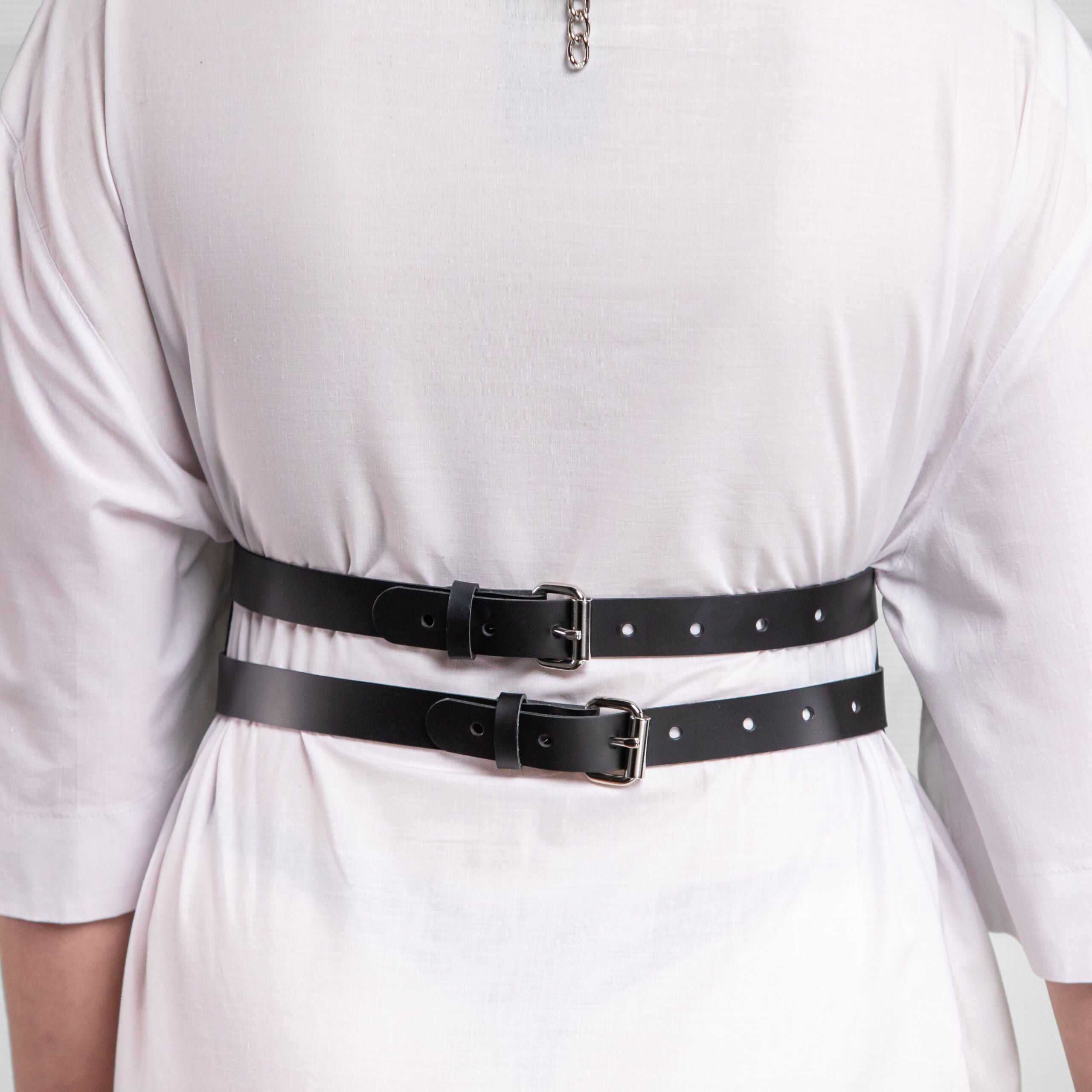 The person wearing a white dress and a black harness or belt with a circular metal ring. They have a mask covering their lower face, featuring a fringe design. Their hair is styled and they have dramatic makeup. The backdrop appears neutral, focusing attention on the person's outfit and accessories.