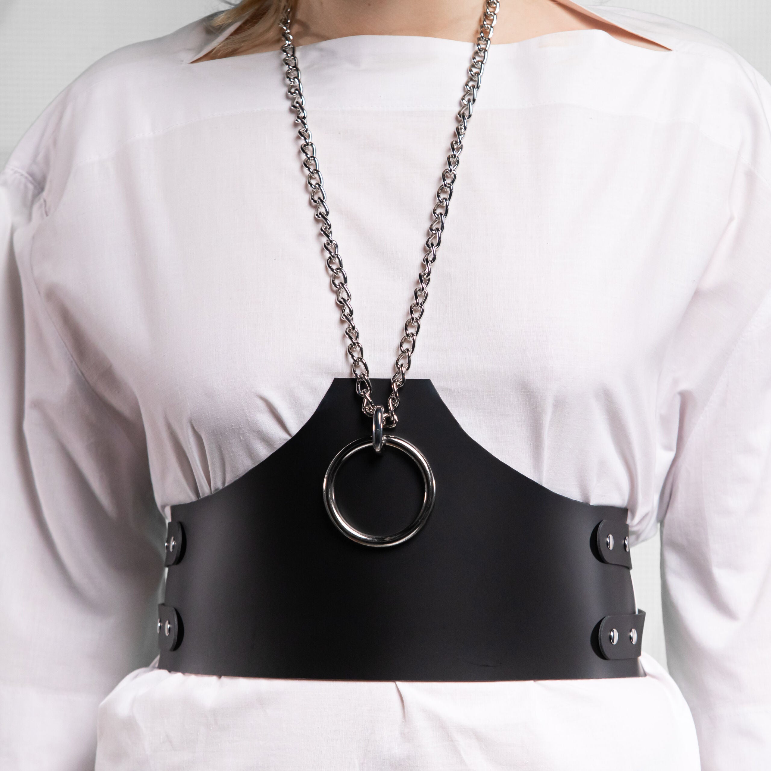 The person wearing a white dress and a black harness or belt with a circular metal ring. They have a mask covering their lower face, featuring a fringe design. Their hair is styled and they have dramatic makeup. The backdrop appears neutral, focusing attention on the person's outfit and accessories.