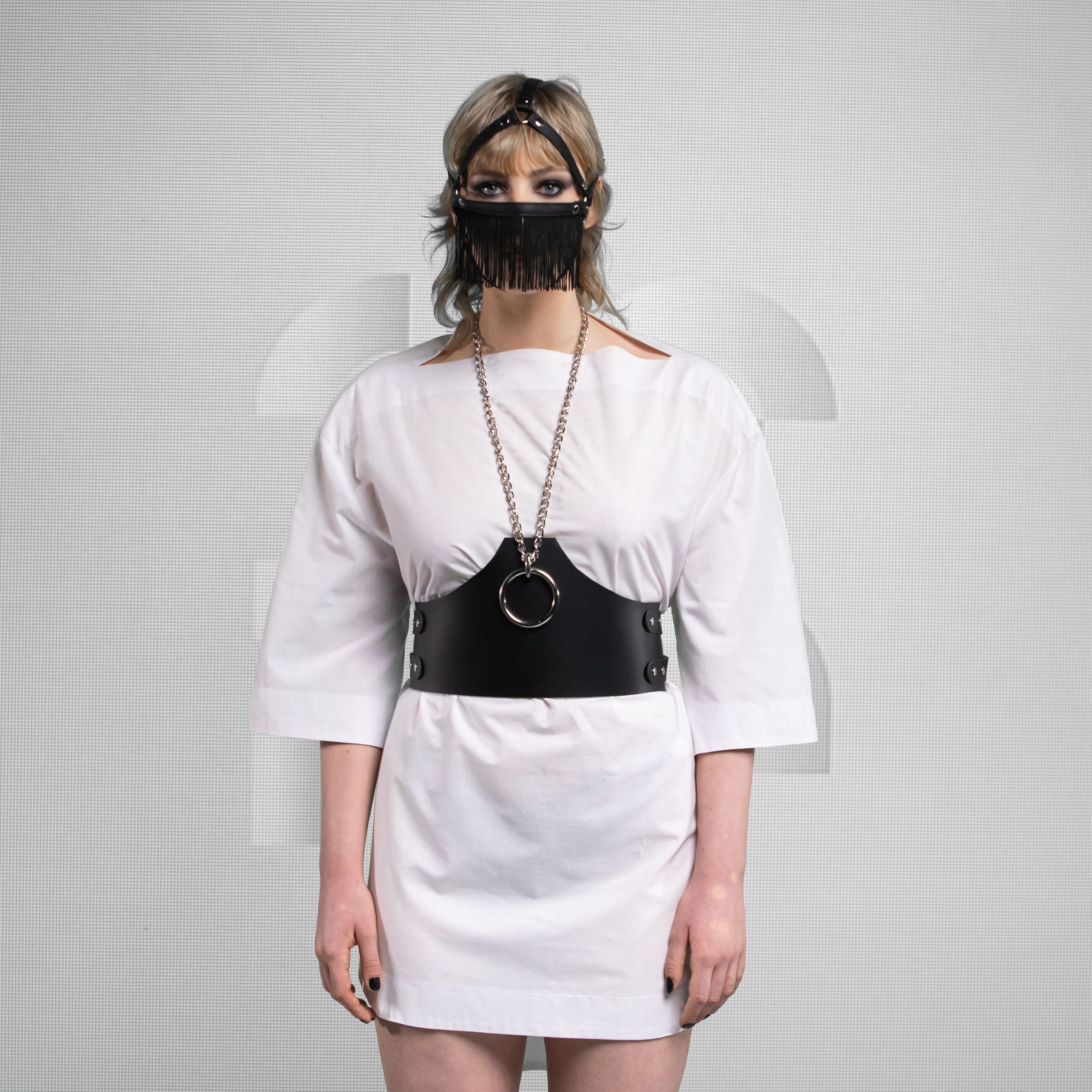 The person wearing a white dress and a black harness or belt with a circular metal ring. They have a mask covering their lower face, featuring a fringe design. Their hair is styled and they have dramatic makeup. The backdrop appears neutral, focusing attention on the person's outfit and accessories.