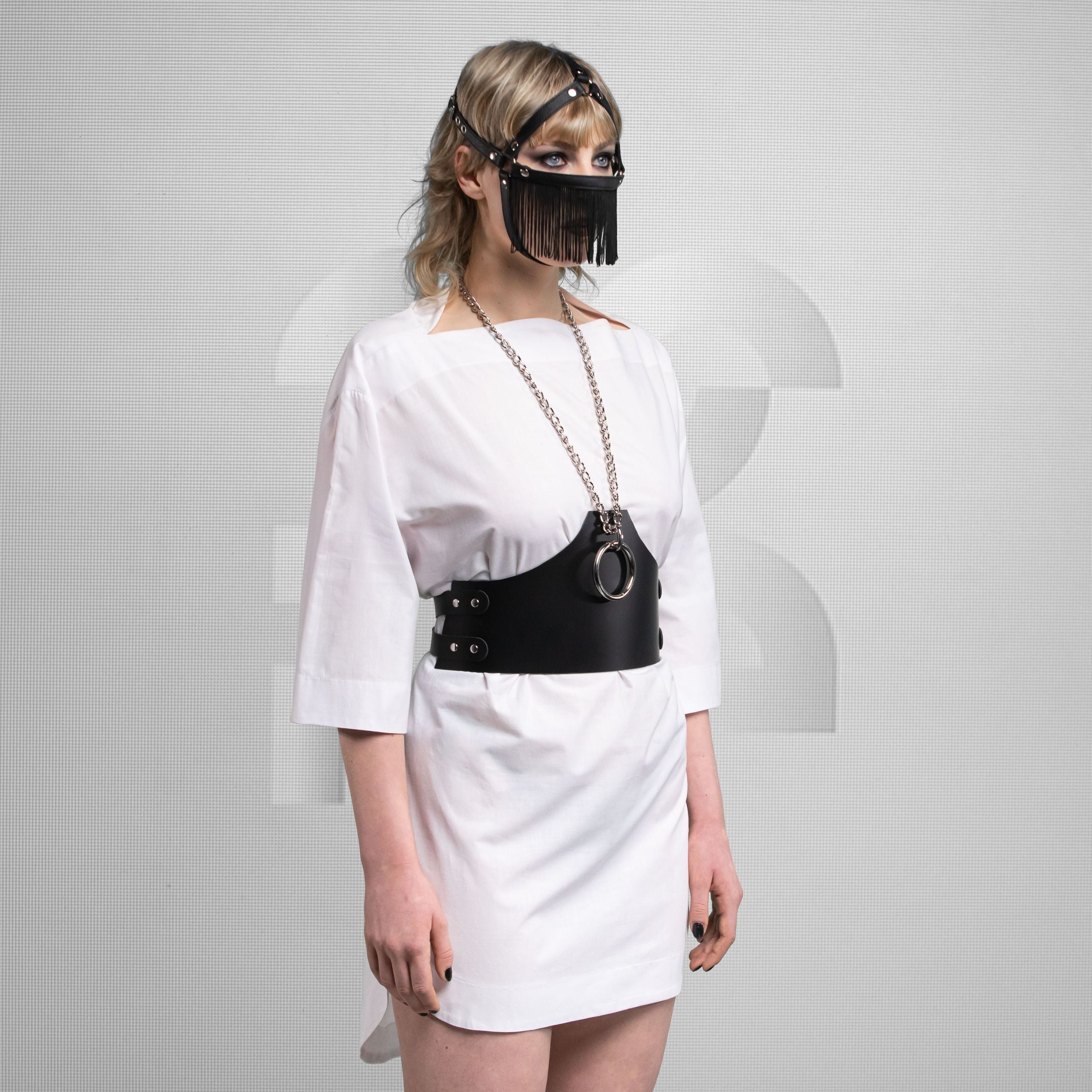The person wearing a white dress and a black harness or belt with a circular metal ring. They have a mask covering their lower face, featuring a fringe design. Their hair is styled and they have dramatic makeup. The backdrop appears neutral, focusing attention on the person's outfit and accessories.