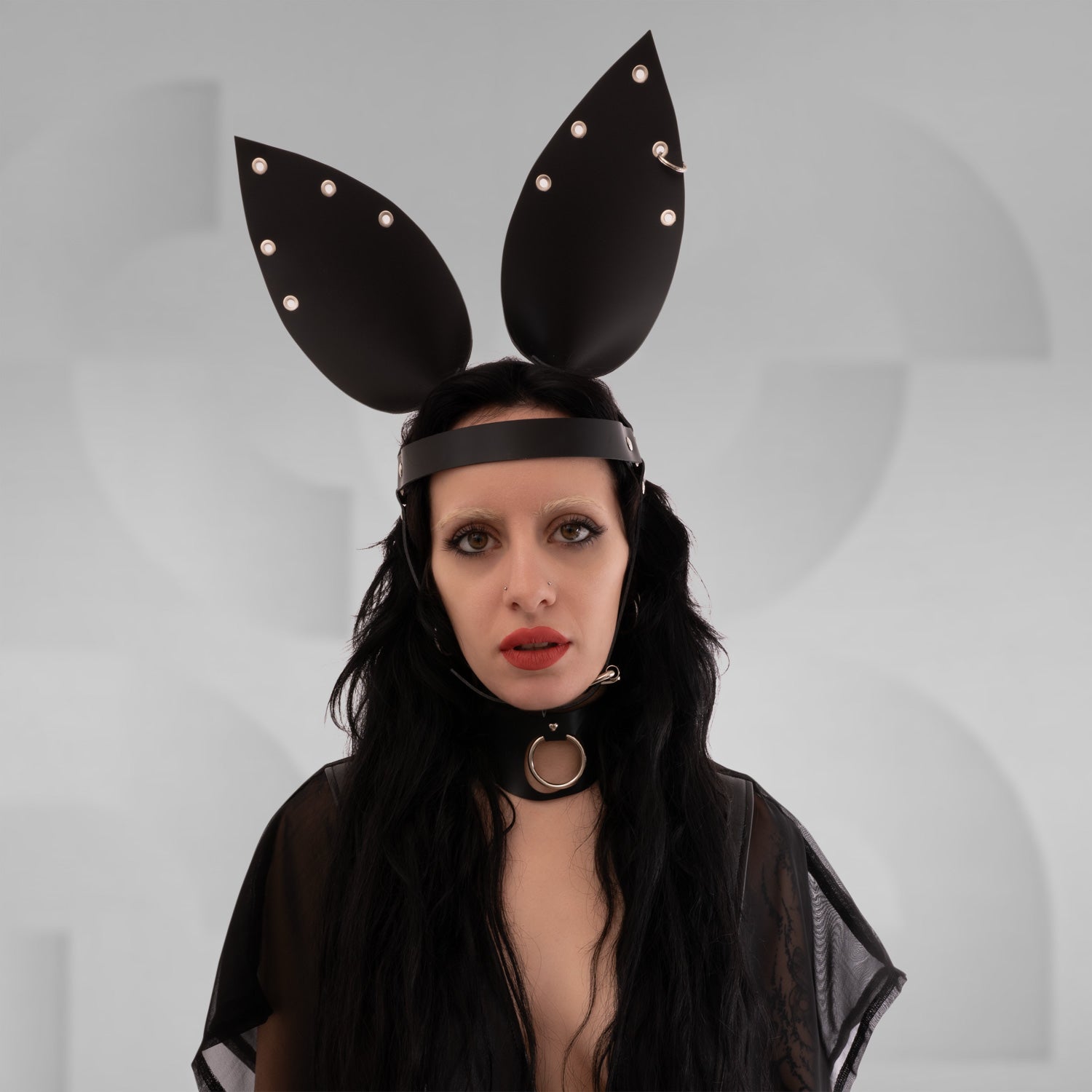 The is facing forward, wearing a similar headpiece with large, pointed extensions that resemble bunny ears, also made of a dark material with metal studs. They have long, dark hair and striking makeup, featuring dark lips and accentuated eyes. A choker with a circular detail is around their neck. The background remains neutral, drawing attention to their unique look and accessories.