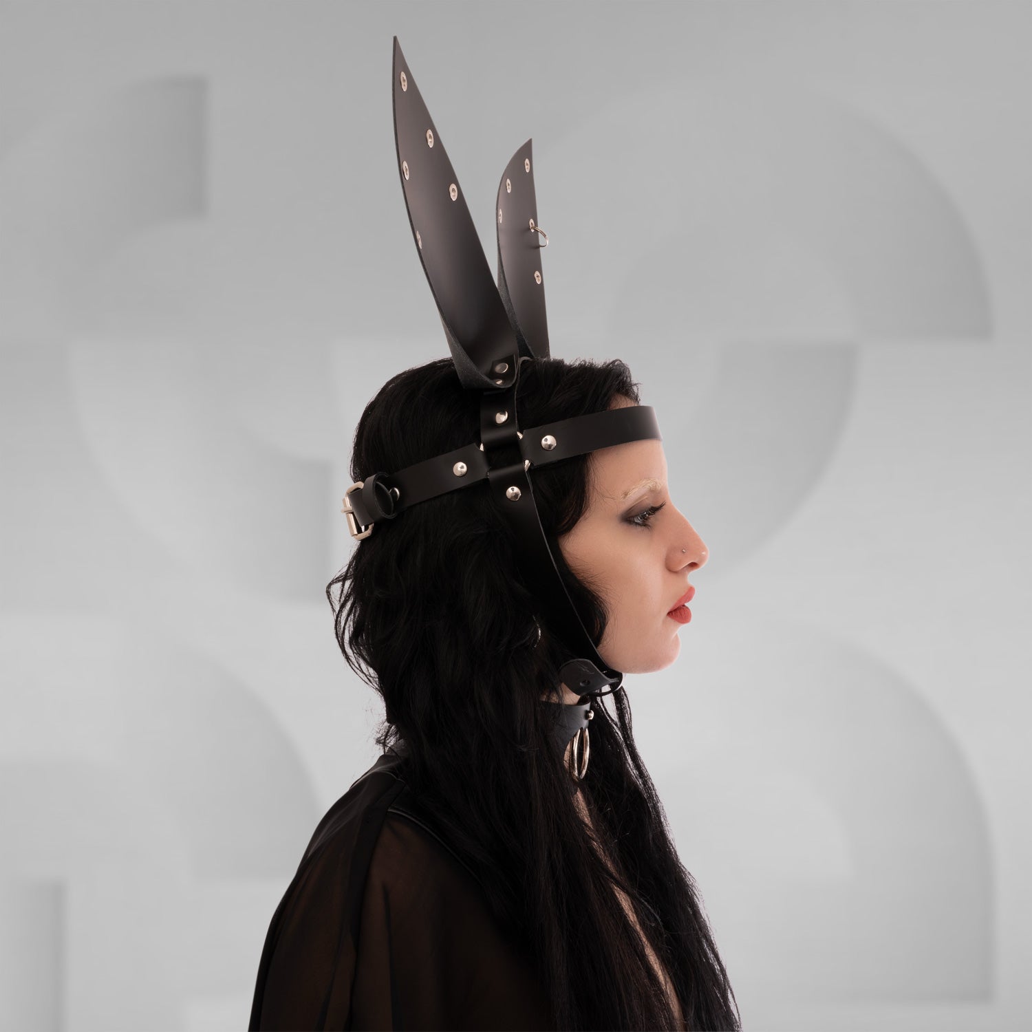The is shown in profile view, wearing a headpiece with tall, vertical extensions resembling ears. The headpiece is made of a dark material with metal rivets. Their hair is dark and long, and they have dramatic makeup with dark eyeliner. The background is neutral, emphasizing the headpiece and styling.