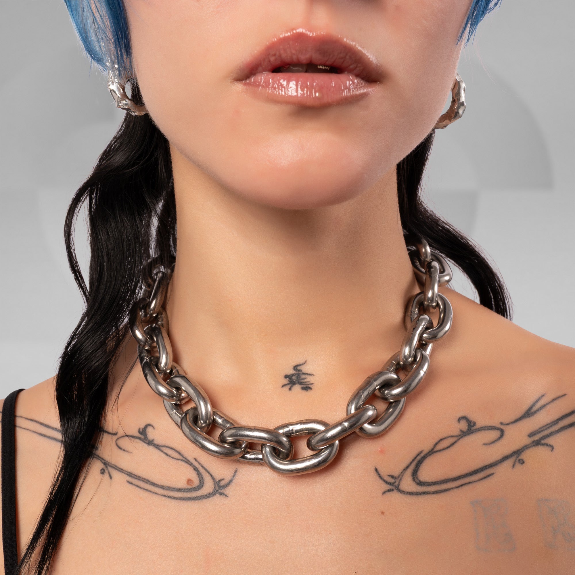 A close-up of a model wearing a chunky, oversized silver chain necklace. The model has blue hair, black hair, and visible tattoos on the chest.