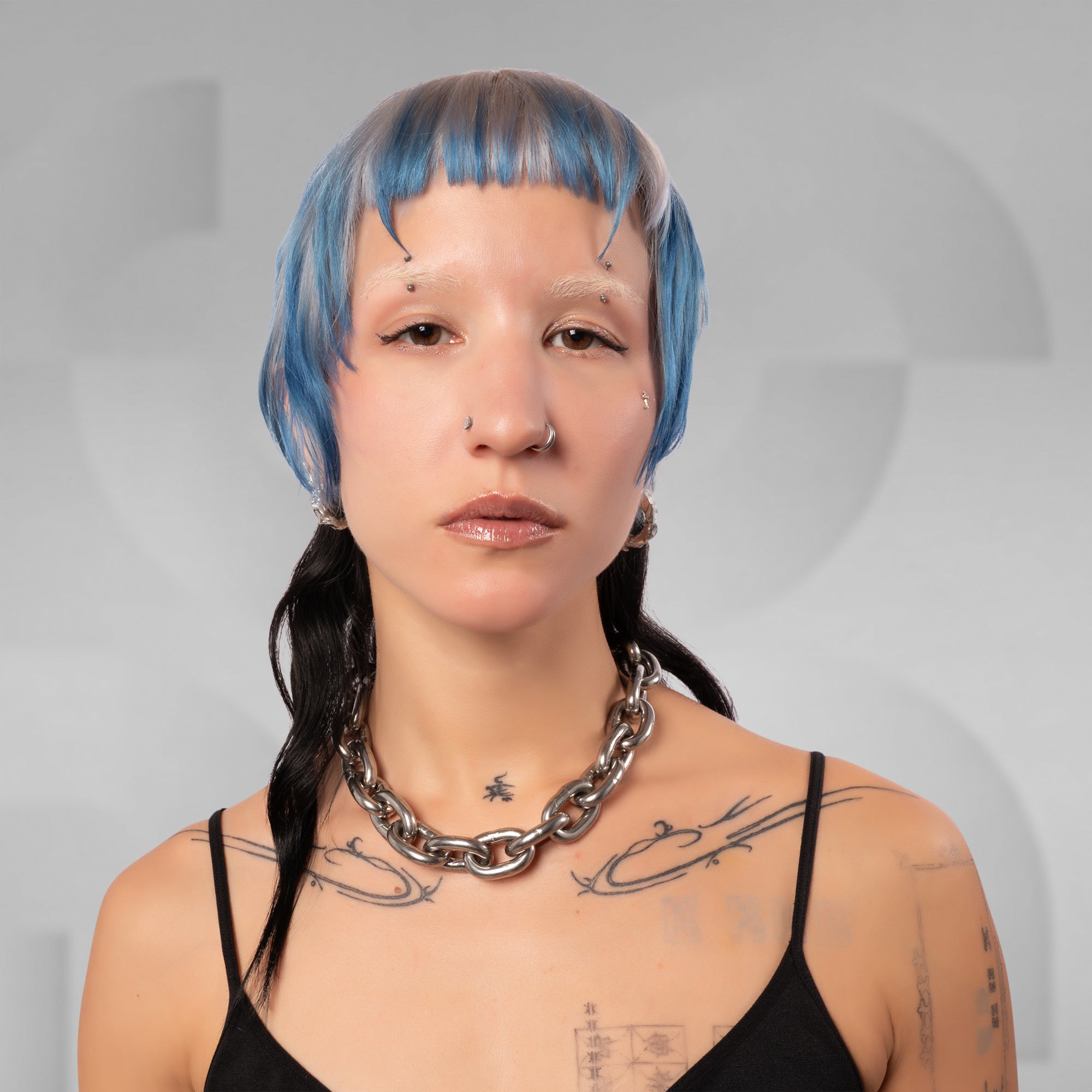 A close-up portrait of a person with short blue hair, a black spaghetti-strap top, and a chunky silver chain necklace against a light gray background.