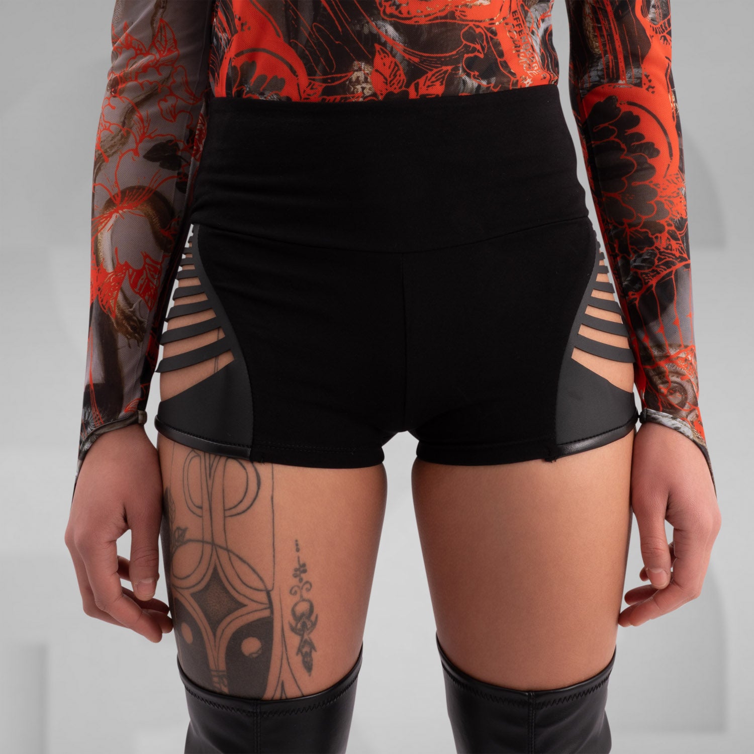 Model wearing high-waisted black hotpants featuring cut-out side details, paired with thigh-high boots and a patterned long-sleeve top, highlighting a bold and fashionable look.