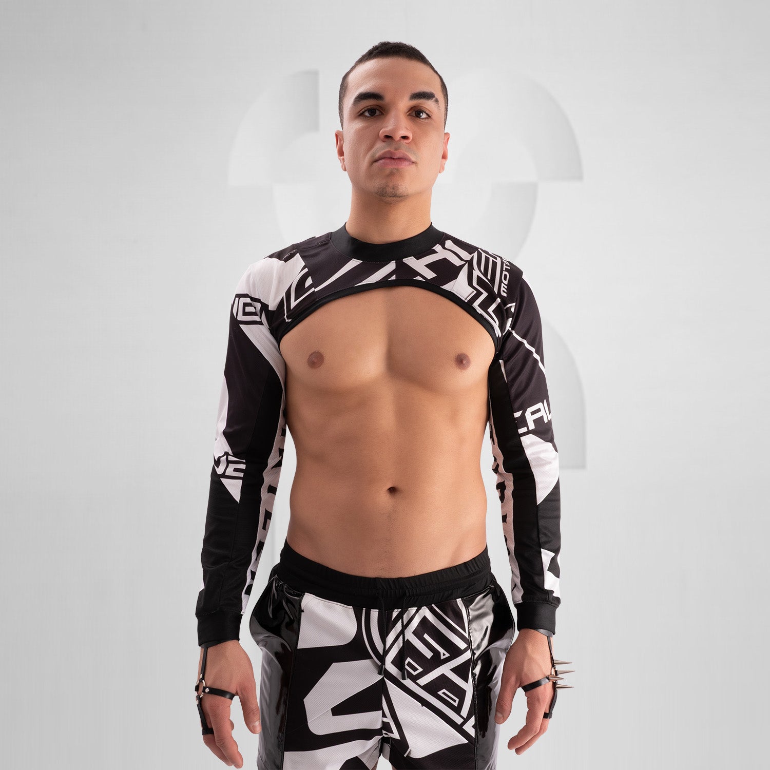 The image showcases a person wearing a bold, geometric outfit with strong black and white patterns. The top is a distinctive design featuring long sleeves and an open front, creating an avant-garde look. The matching pants continue the geometric theme. The overall style is modern and edgy, reflecting influences from both high fashion and the techno music scene.