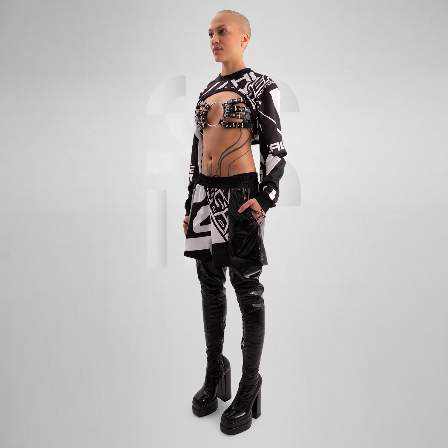 The image showcases a person wearing a bold, geometric outfit with strong black and white patterns. The top is a distinctive design featuring long sleeves and an open front, creating an avant-garde look. The matching pants continue the geometric theme. The overall style is modern and edgy, reflecting influences from both high fashion and the techno music scene.