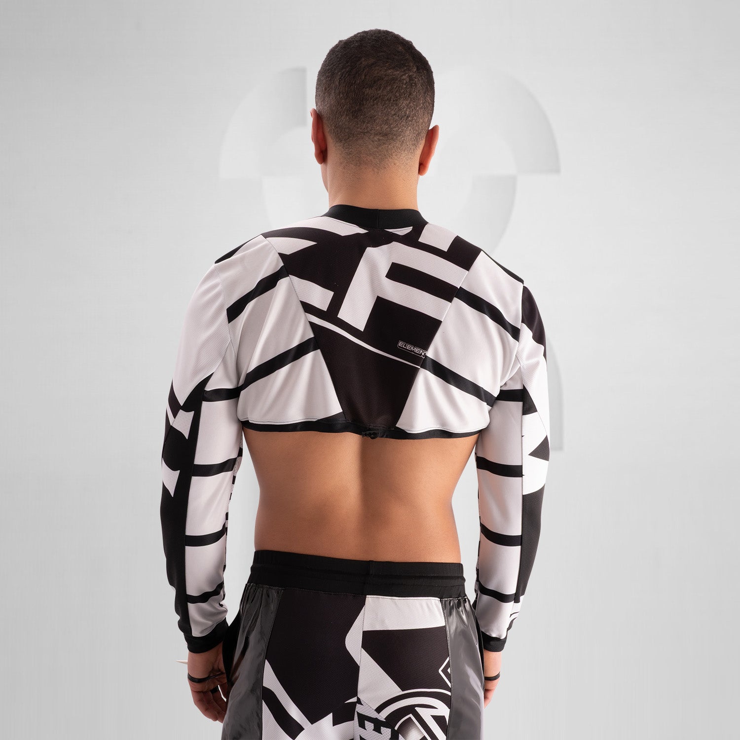 The image showcases a person wearing a bold, geometric outfit with strong black and white patterns. The top is a distinctive design featuring long sleeves and an open front, creating an avant-garde look. The matching pants continue the geometric theme. The overall style is modern and edgy, reflecting influences from both high fashion and the techno music scene.