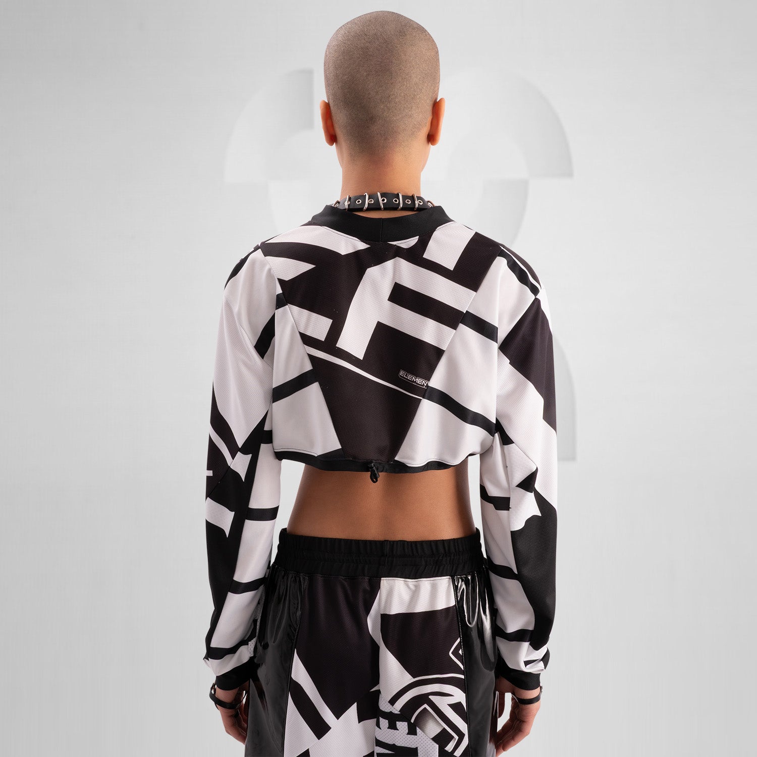 The image showcases a person wearing a bold, geometric outfit with strong black and white patterns. The top is a distinctive design featuring long sleeves and an open front, creating an avant-garde look. The matching pants continue the geometric theme. The overall style is modern and edgy, reflecting influences from both high fashion and the techno music scene.
