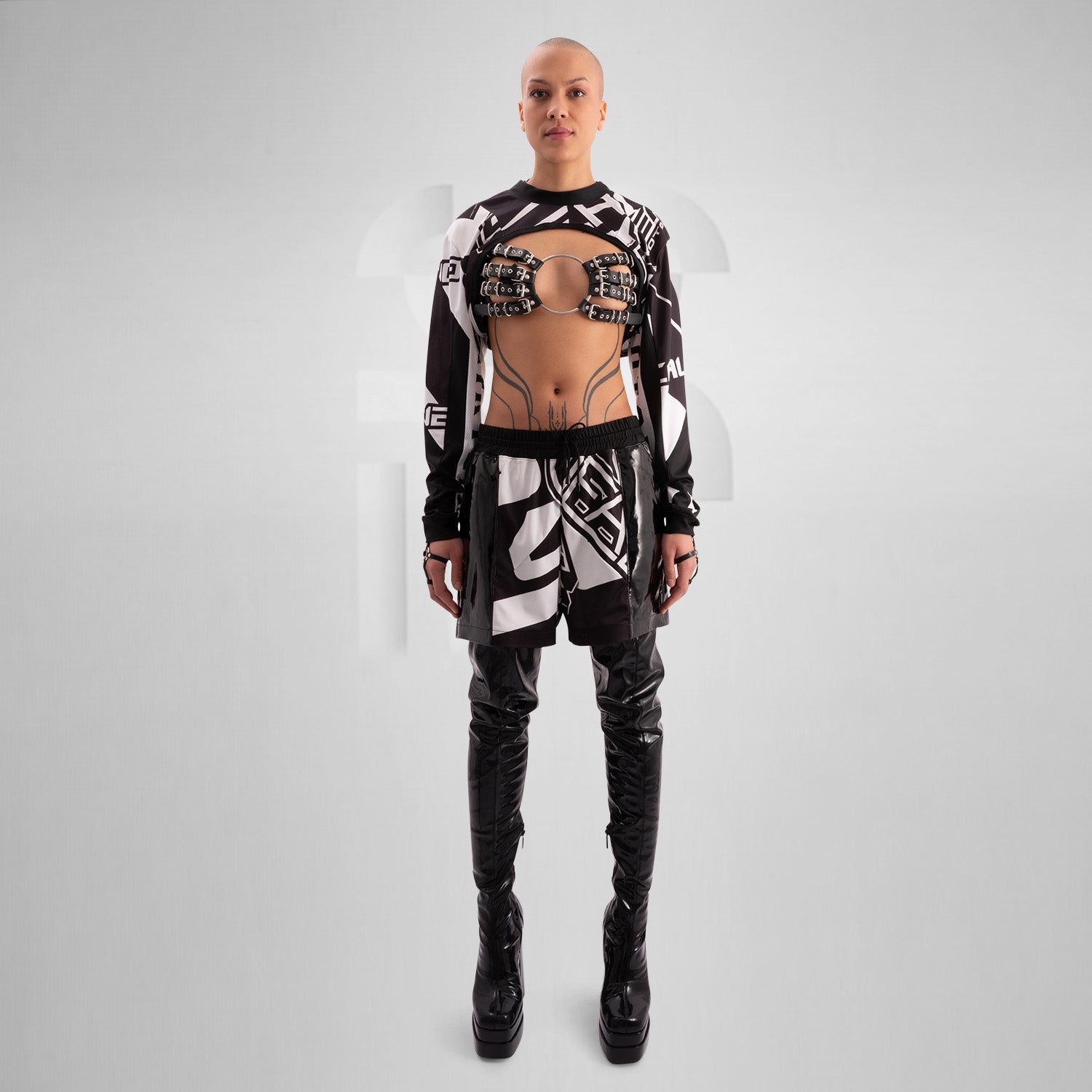 The image showcases a person wearing a bold, geometric outfit with strong black and white patterns. The top is a distinctive design featuring long sleeves and an open front, creating an avant-garde look. The matching pants continue the geometric theme. The overall style is modern and edgy, reflecting influences from both high fashion and the techno music scene.