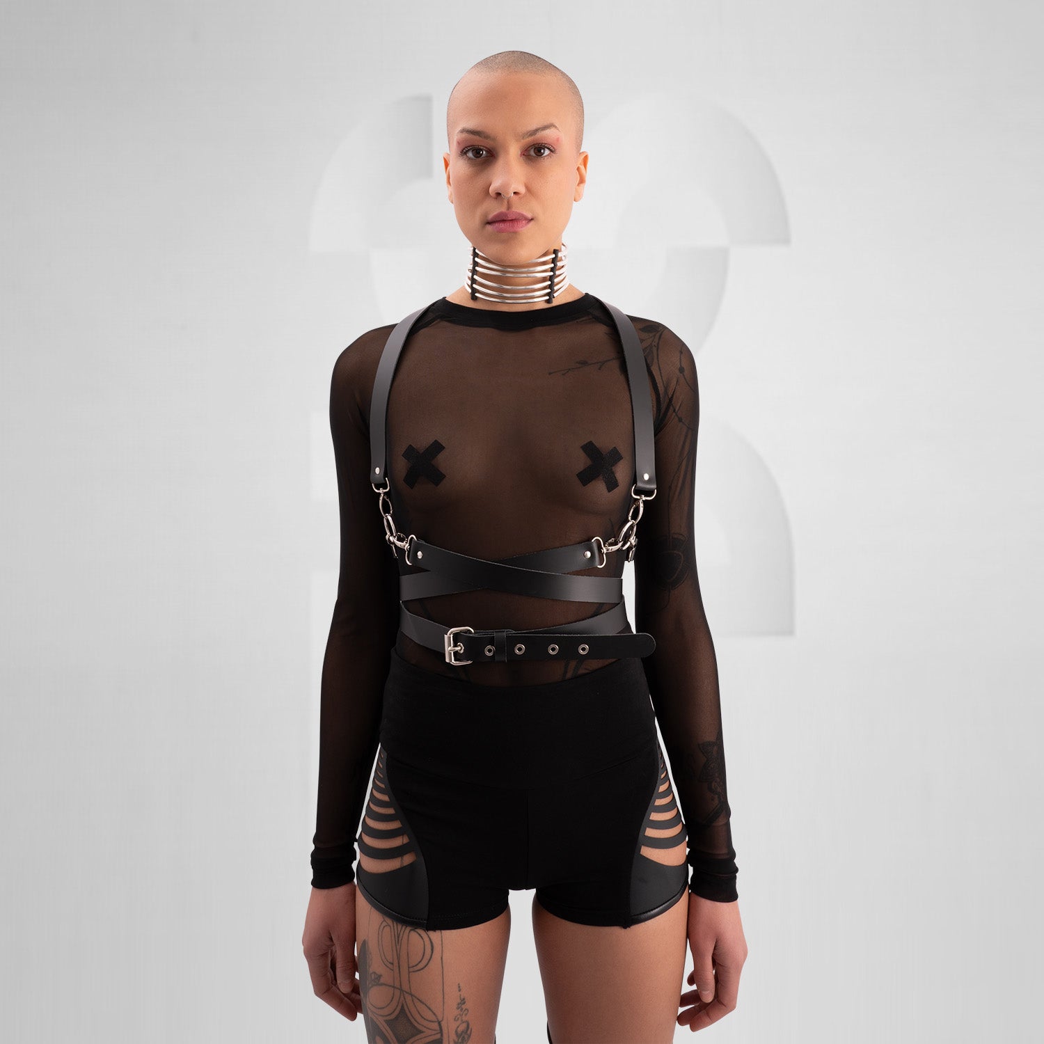 "Bold, avant-garde outfit featuring a black leather harness with silver hardware, a sheer long-sleeve top with X-shaped pasties, and edgy black hotpants with laser-cut vegan leather details. The model exudes confidence with a shaved head, statement choker, and intricate tattoos, set against a minimalistic background."