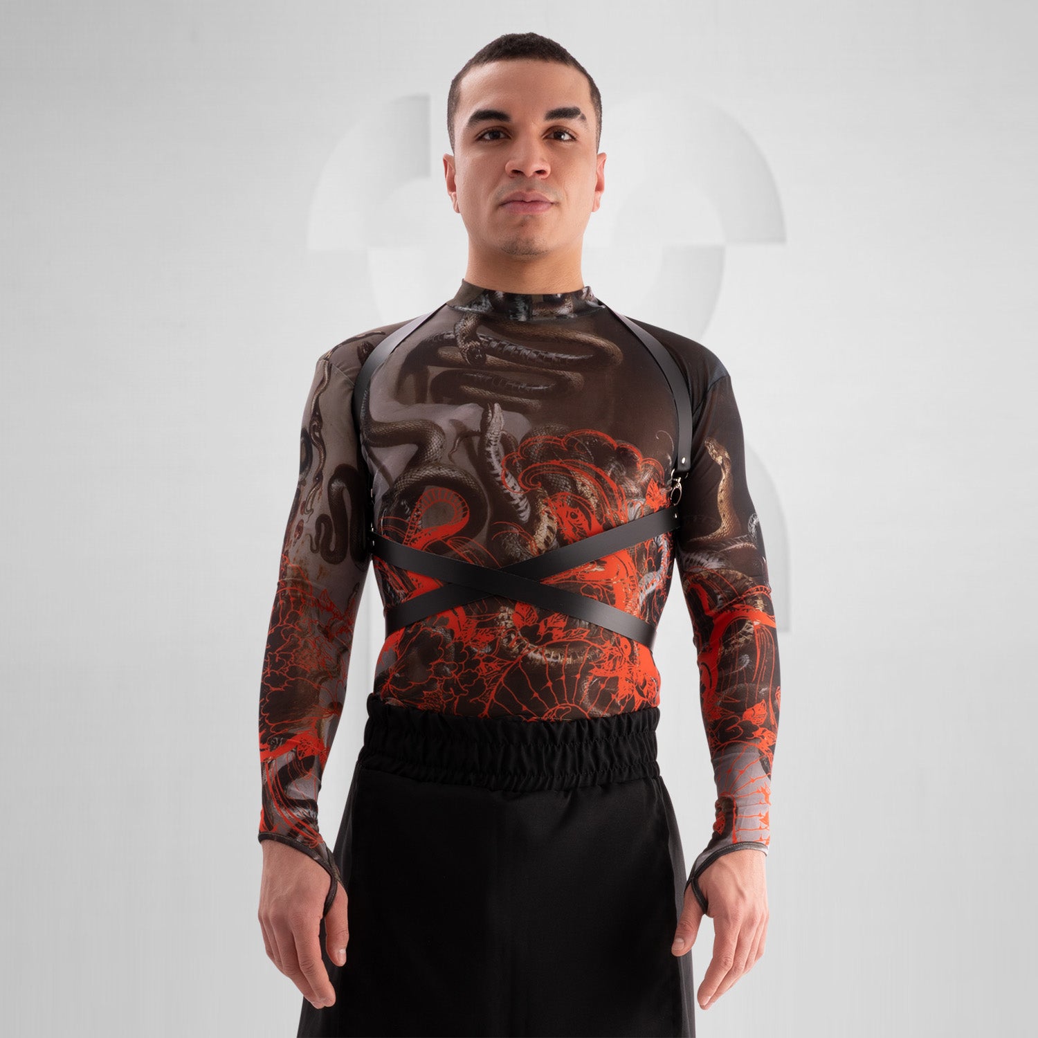 "Bold, avant-garde outfit featuring a black leather harness with silver hardware, a sheer long-sleeve top with X-shaped pasties, and edgy black hotpants with laser-cut vegan leather details. The model exudes confidence with a shaved head, statement choker, and intricate tattoos, set against a minimalistic background."