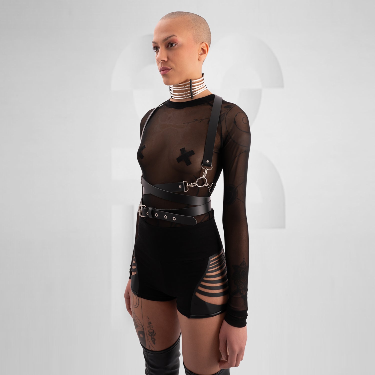 "Bold, avant-garde outfit featuring a black leather harness with silver hardware, a sheer long-sleeve top with X-shaped pasties, and edgy black hotpants with laser-cut vegan leather details. The model exudes confidence with a shaved head, statement choker, and intricate tattoos, set against a minimalistic background."