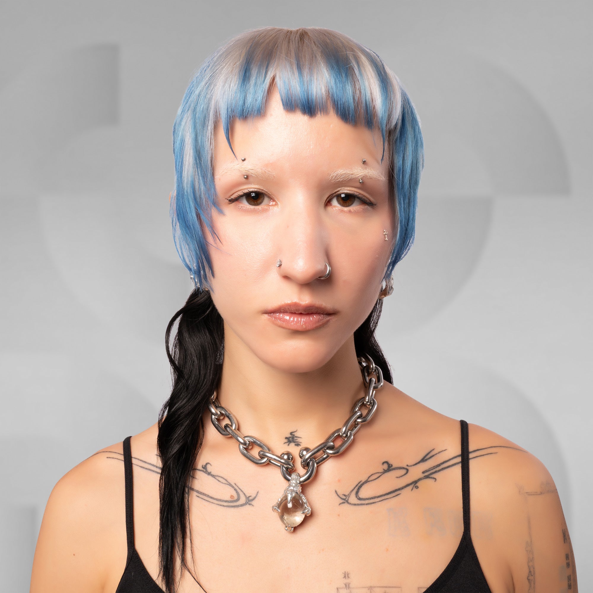A close-up portrait of a person with a short blue and gray wig, black braided hair, silver nose jewelry, and a chunky silver chain necklace, wearing a black spaghetti-strap top against a light gray background.
