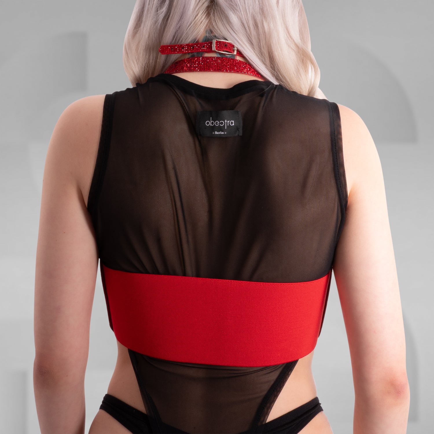 This image shows a person wearing a fashion outfit that includes a sheer black top and a red harness or corset-style accessory with buckle details. The accessory is made of a textured, glittery material. Additionally, the person is wearing a matching red choker. This style is edgy and modern, often seen in alternative fashion scenes.