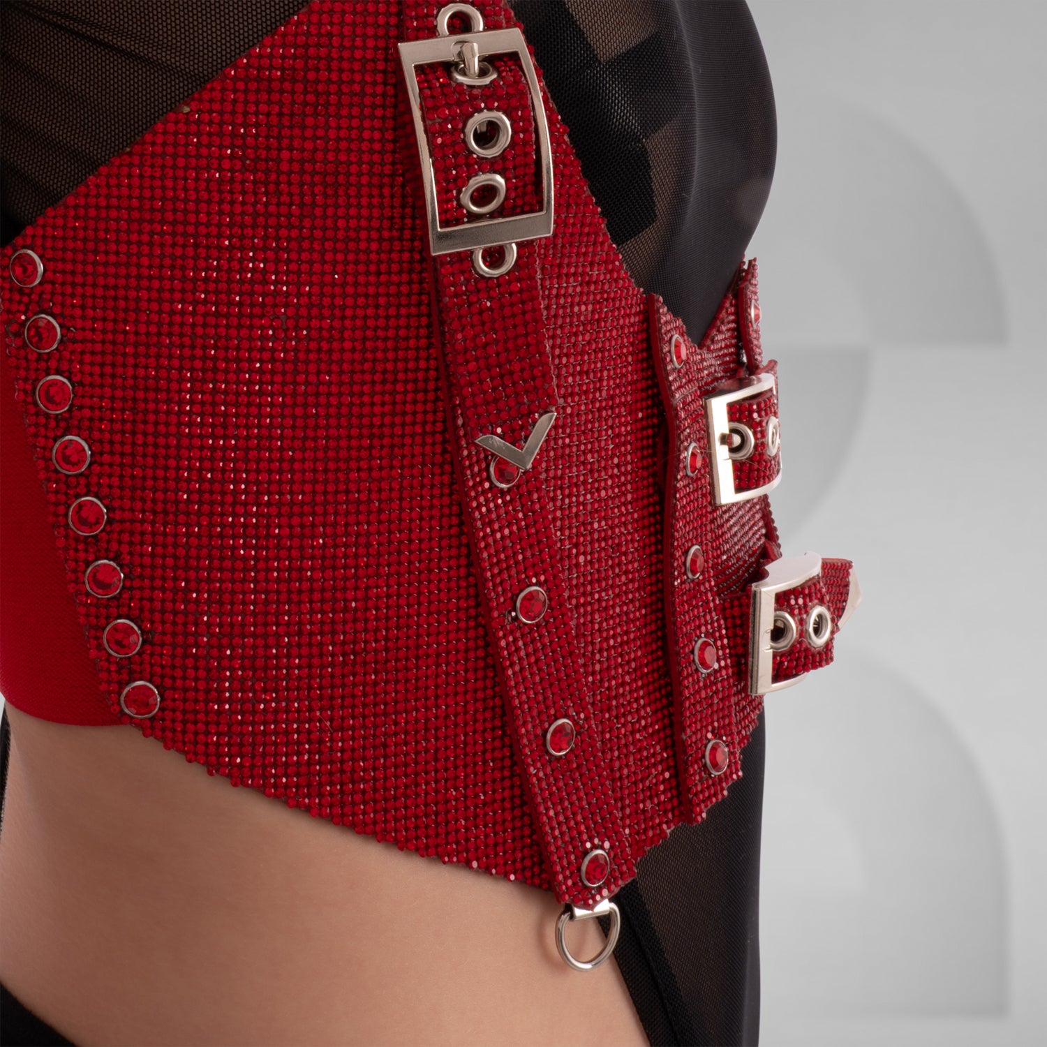 This image shows a person wearing a fashion outfit that includes a sheer black top and a red harness or corset-style accessory with buckle details. The accessory is made of a textured, glittery material. Additionally, the person is wearing a matching red choker. This style is edgy and modern, often seen in alternative fashion scenes.