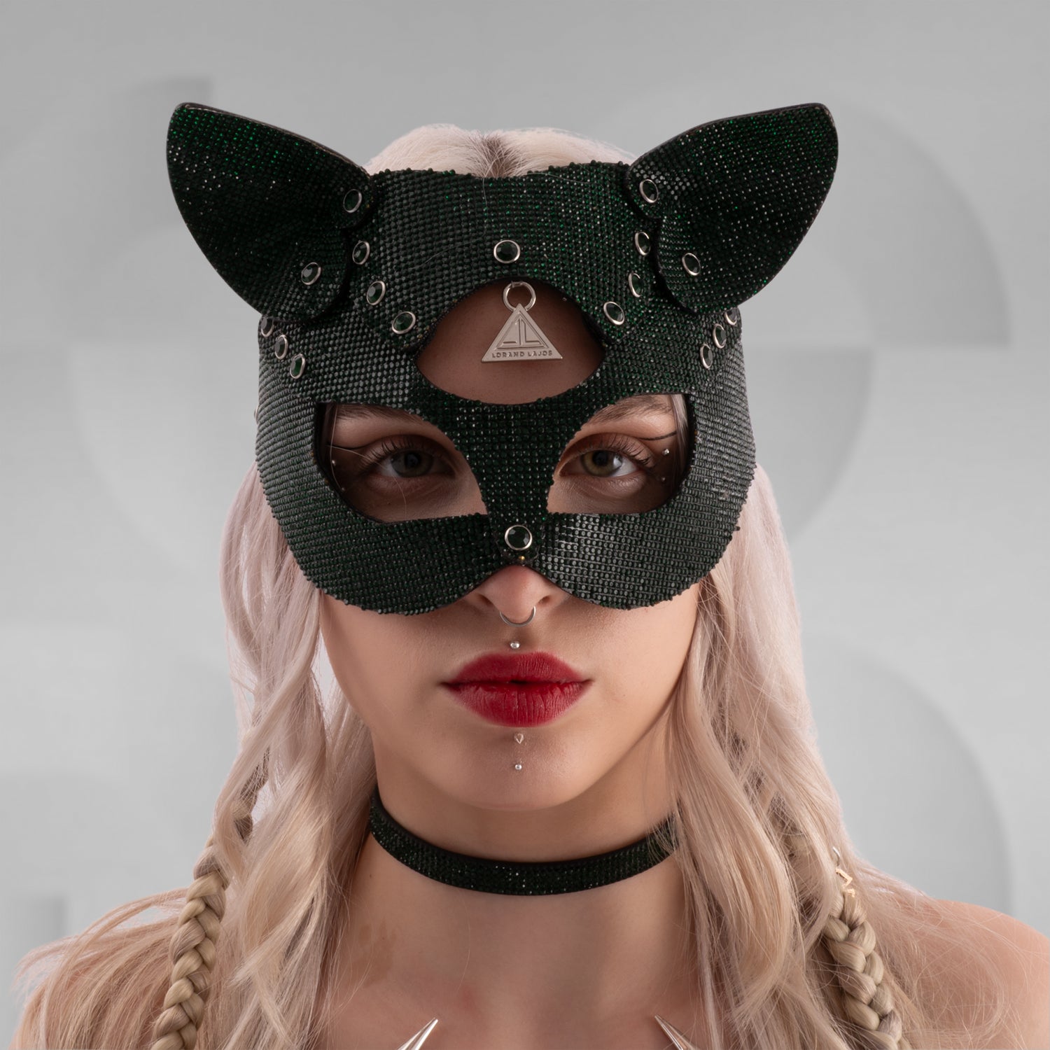 Woman wearing a sparkling rhinestone cat mask with pointed ears, paired with a matching crystal choker and shimmering outfit; styled with platinum blonde hair in braids and bold red lipstick, set against a minimalist grey background.