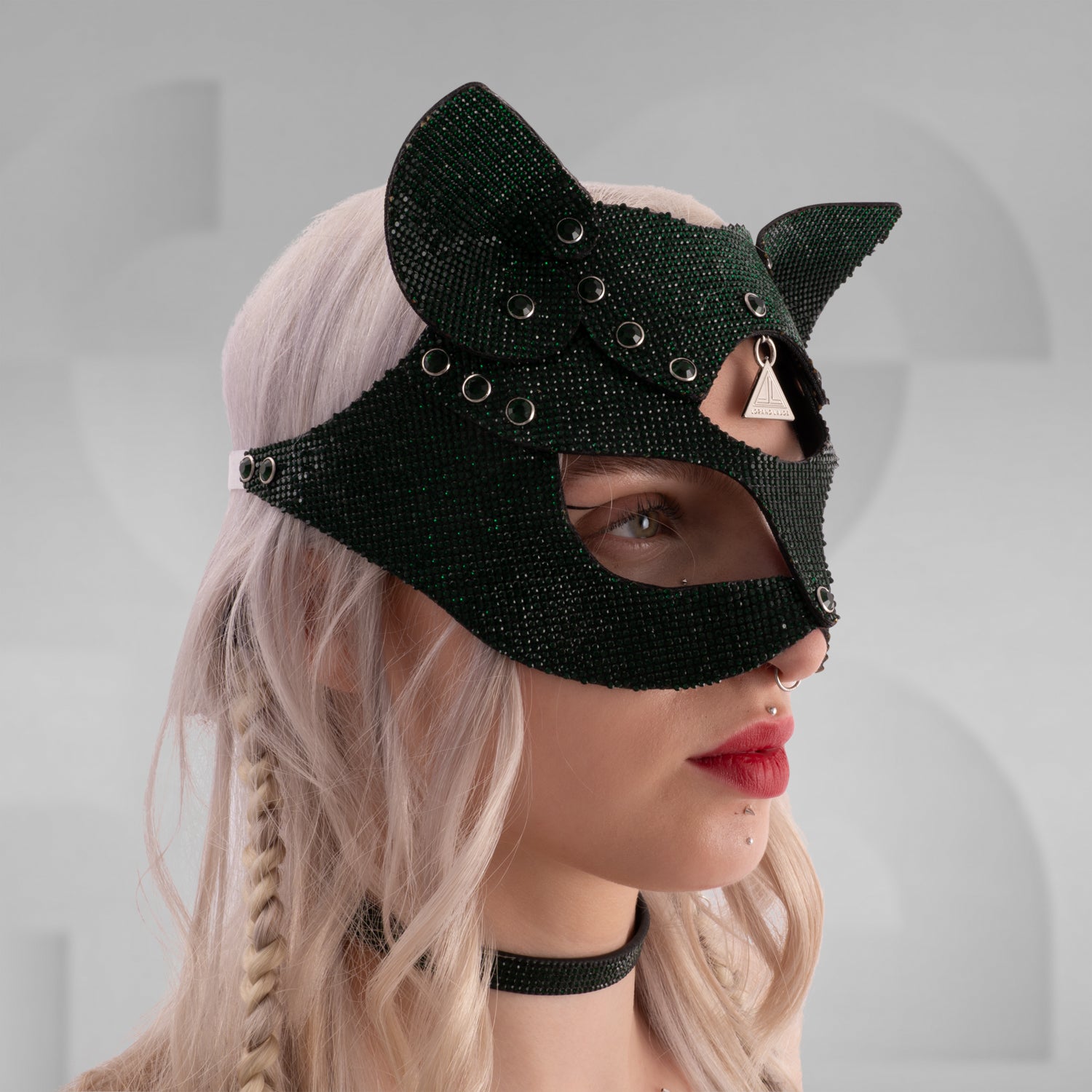 Woman wearing a sparkling rhinestone cat mask with pointed ears, paired with a matching crystal choker and shimmering outfit; styled with platinum blonde hair in braids and bold red lipstick, set against a minimalist grey background.