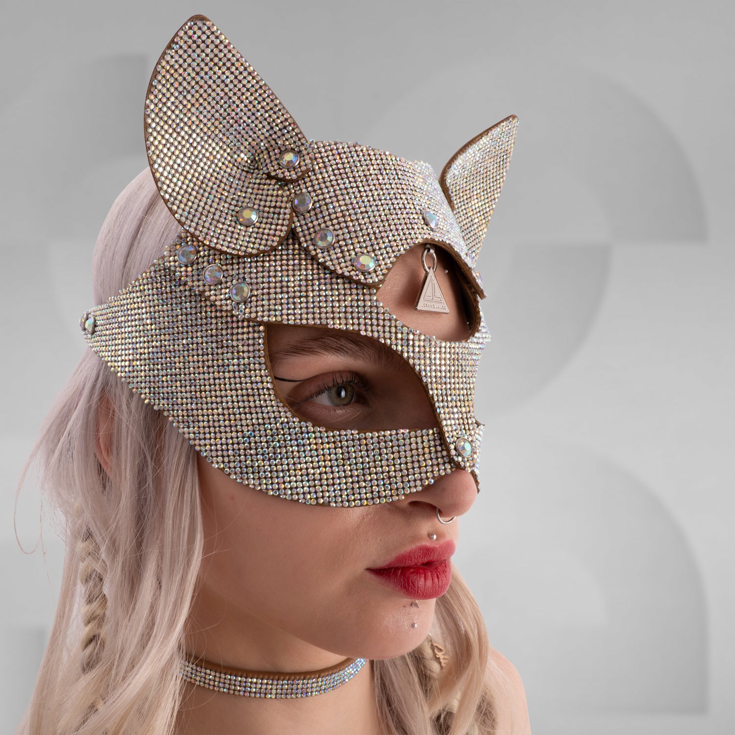 Woman wearing a sparkling rhinestone cat mask with pointed ears, paired with a matching crystal choker and shimmering outfit; styled with platinum blonde hair in braids and bold red lipstick, set against a minimalist grey background.