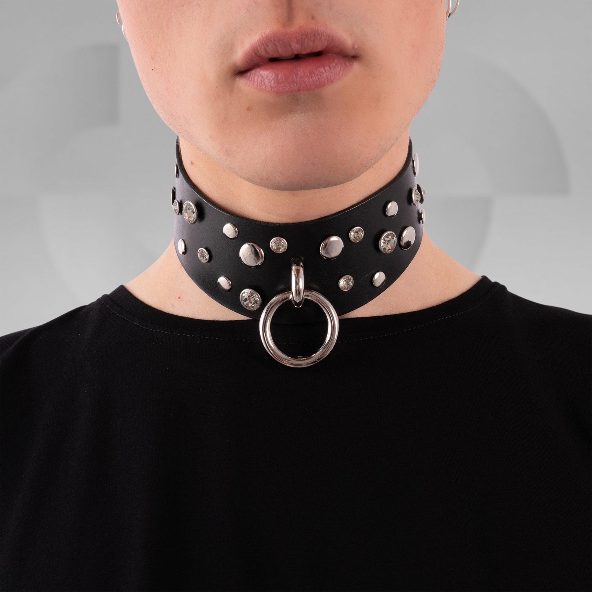 Black leather choker with silver studs and a large ring worn by a person on a gray background