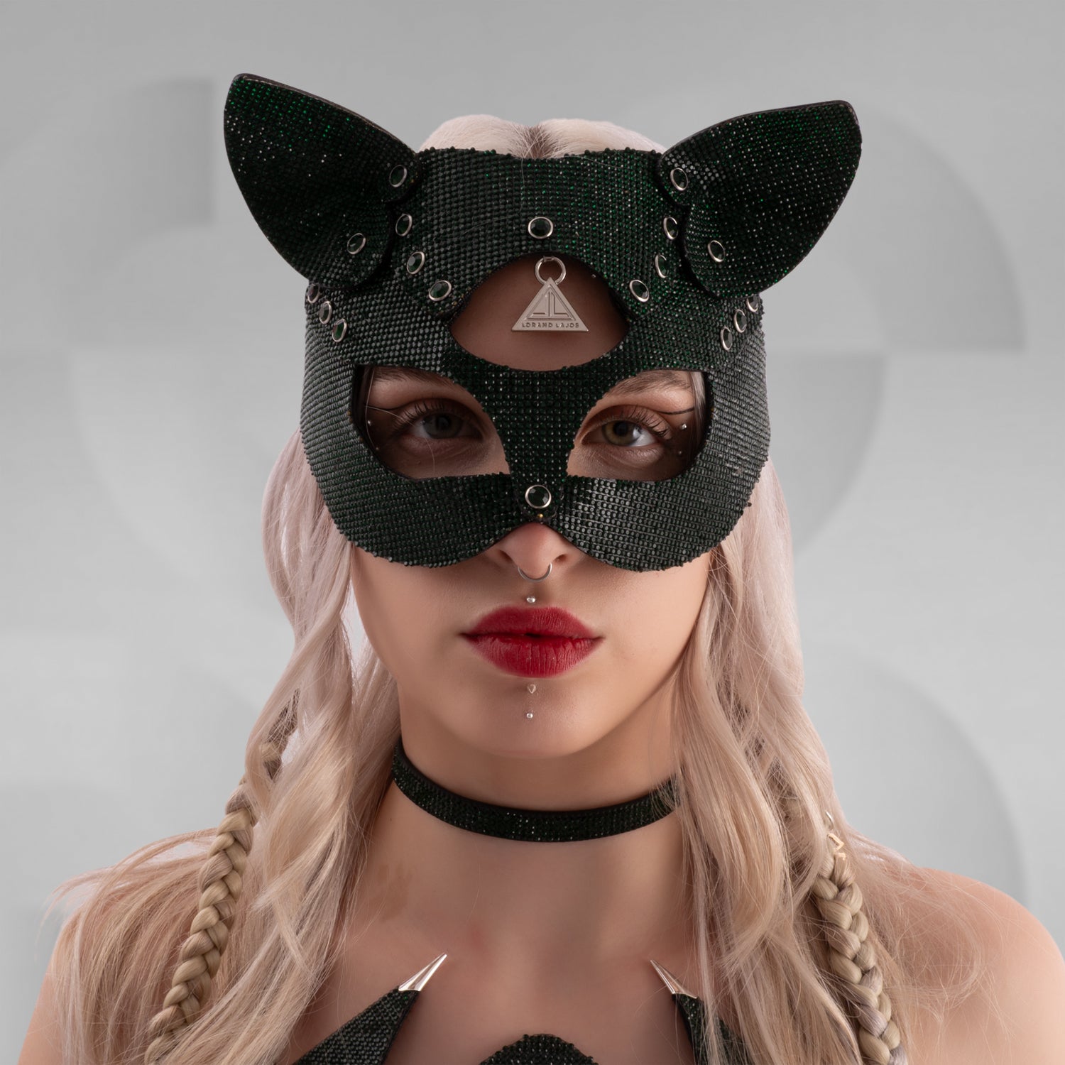 In this picture, you can see a close-up of a woman wearing a striking green rhinestone choker around her neck, which adds a glamorous and edgy vibe. She has platinum blonde hair styled with soft waves, and a small braid on one side. Her makeup features bold red lipstick that matches her choker, and she has a septum piercing as well as two dermal piercings below her lips, adding to her alternative, fashionable style.