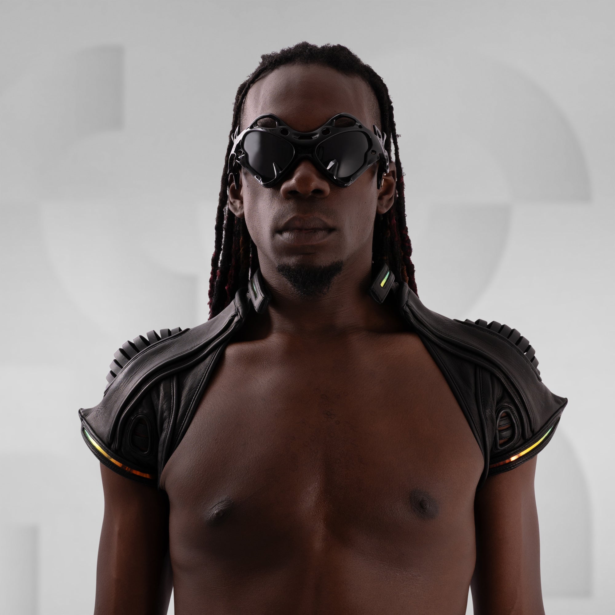 Man wearing futuristic shoulder pads and sunglasses against a gray background