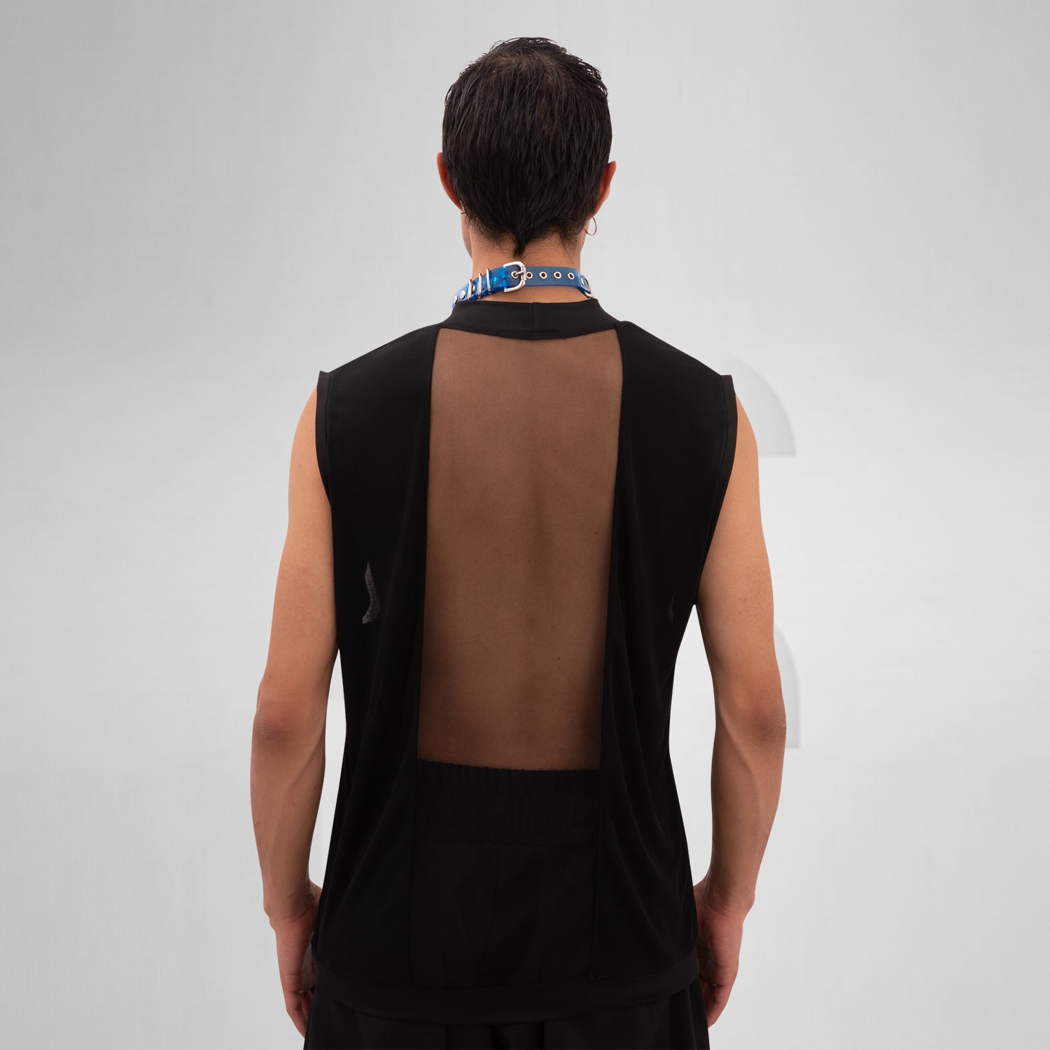 A male model is wearing a black sleeveless tank top with a bold mesh panel across the chest, revealing part of the upper torso. The tank top has a high neckline and a minimalist, modern design, paired with a chunky silver chain necklace. The background is plain and minimal, focusing attention on the garment's details.