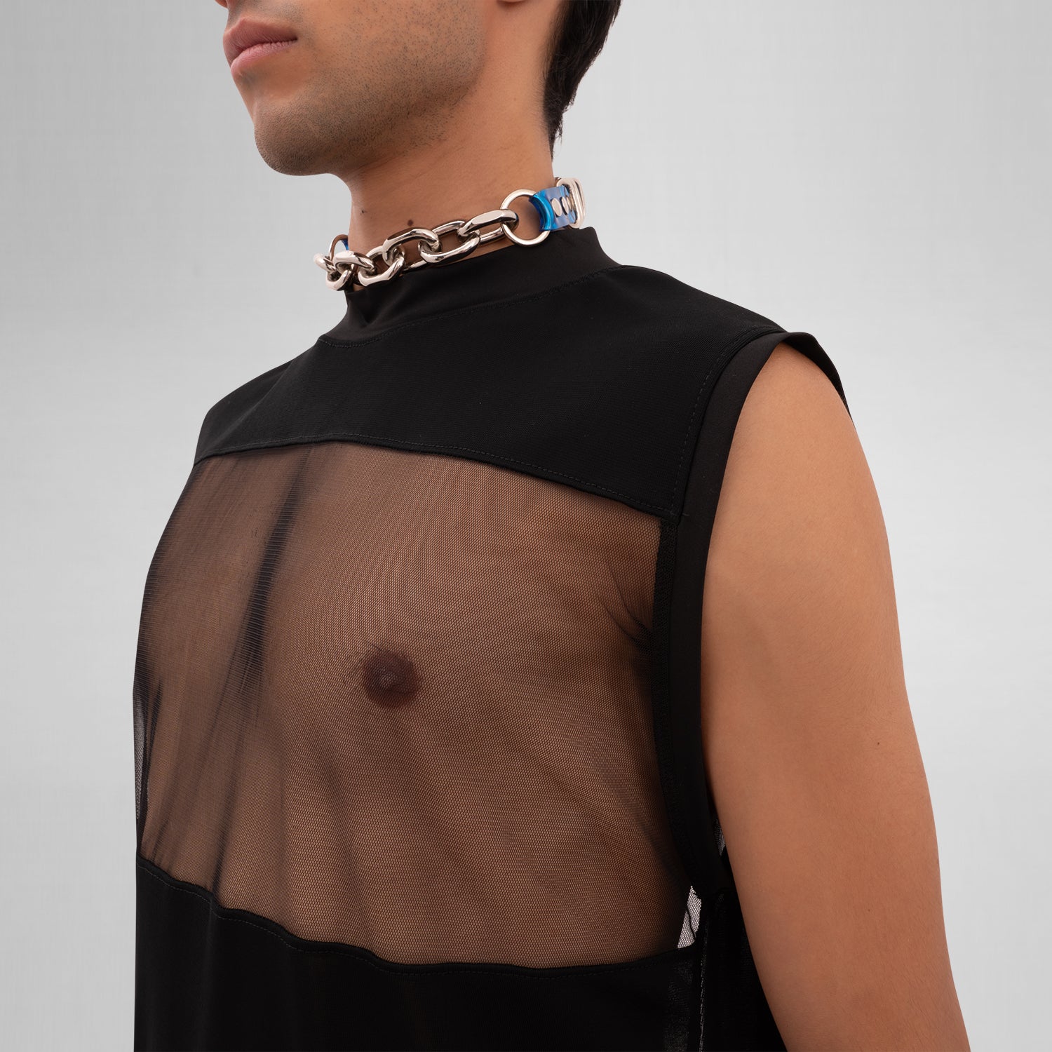 A male model is wearing a black sleeveless tank top with a bold mesh panel across the chest, revealing part of the upper torso. The tank top has a high neckline and a minimalist, modern design, paired with a chunky silver chain necklace. The background is plain and minimal, focusing attention on the garment's details.
