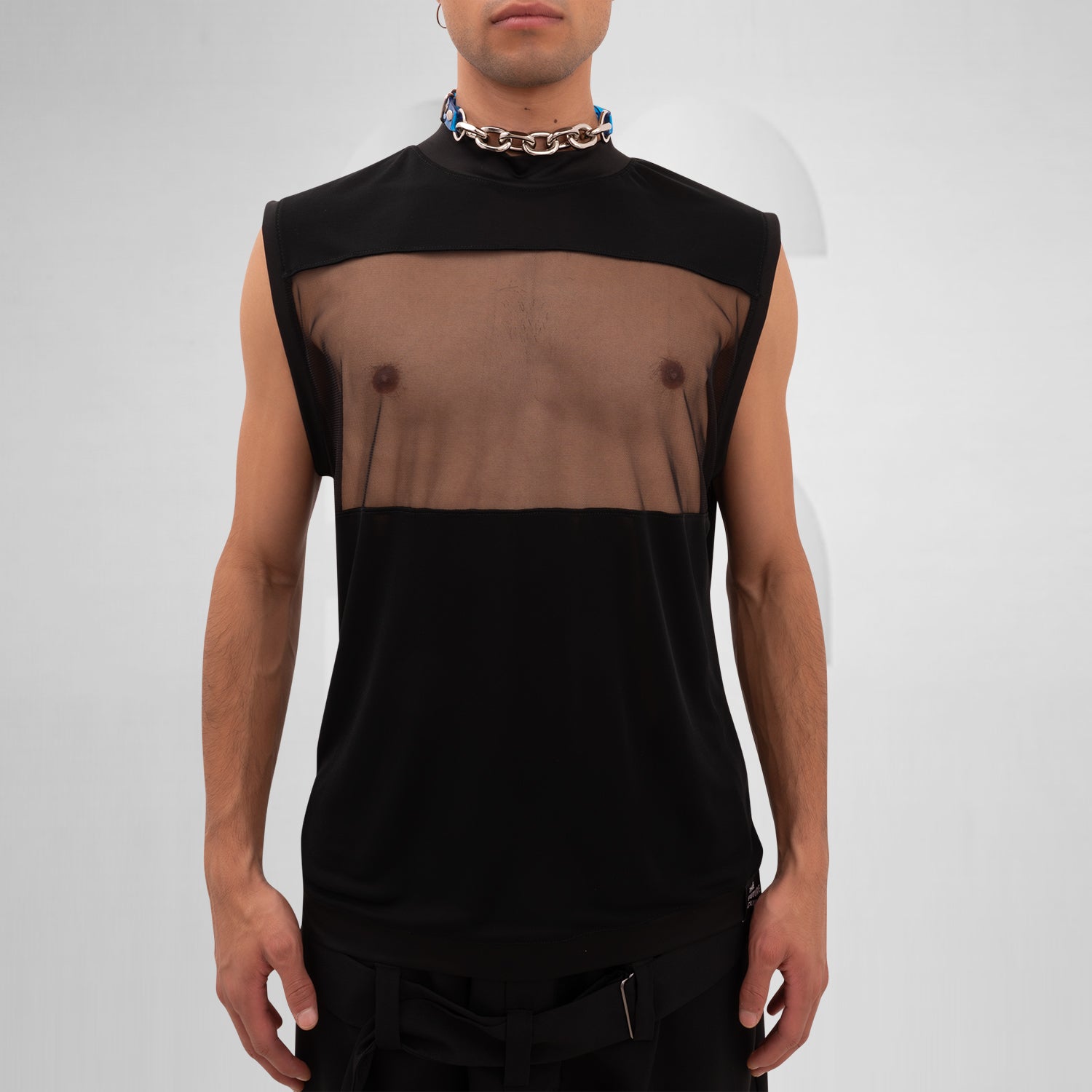 A male model is wearing a black sleeveless tank top with a bold mesh panel across the chest, revealing part of the upper torso. The tank top has a high neckline and a minimalist, modern design, paired with a chunky silver chain necklace. The background is plain and minimal, focusing attention on the garment's details.