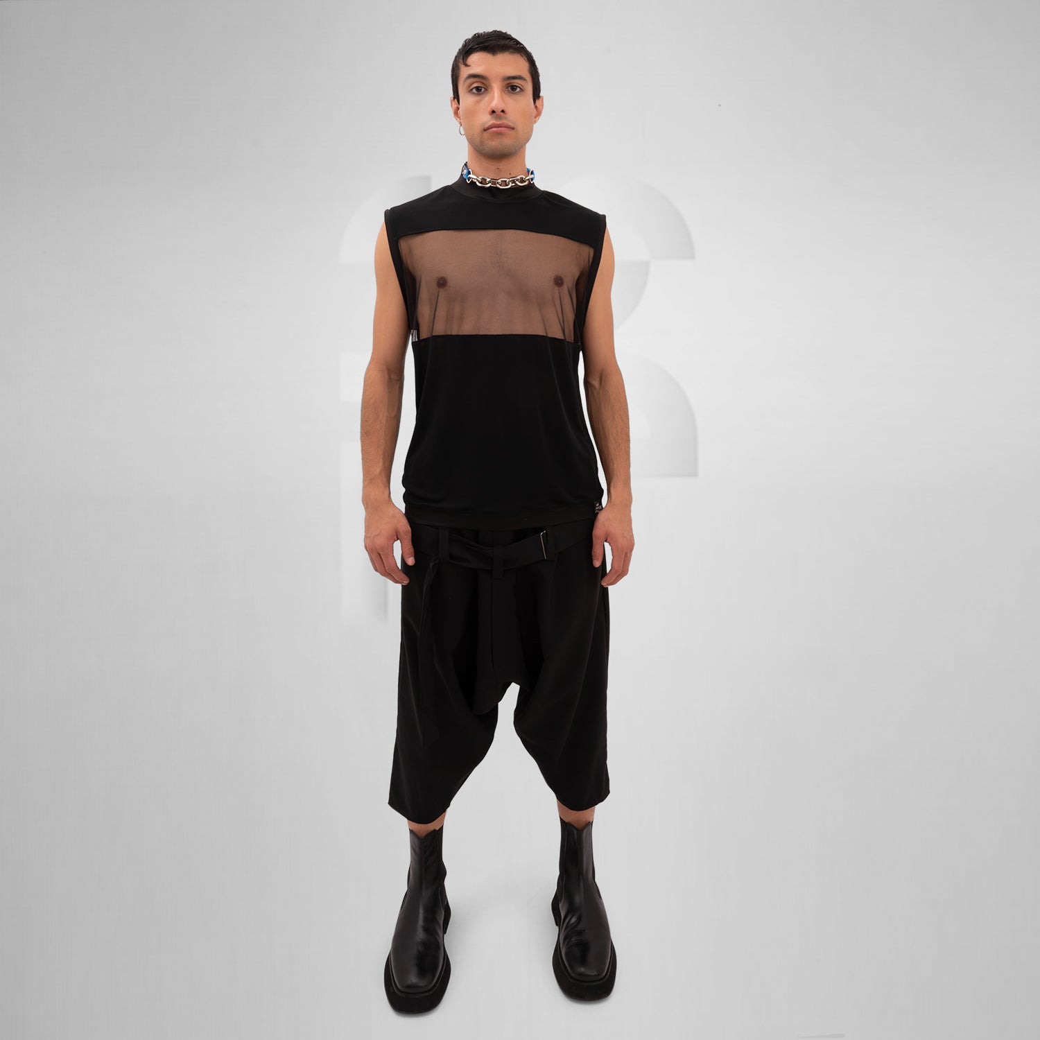 A male model is wearing a black sleeveless tank top with a bold mesh panel across the chest, revealing part of the upper torso. The tank top has a high neckline and a minimalist, modern design, paired with a chunky silver chain necklace. The background is plain and minimal, focusing attention on the garment's details.