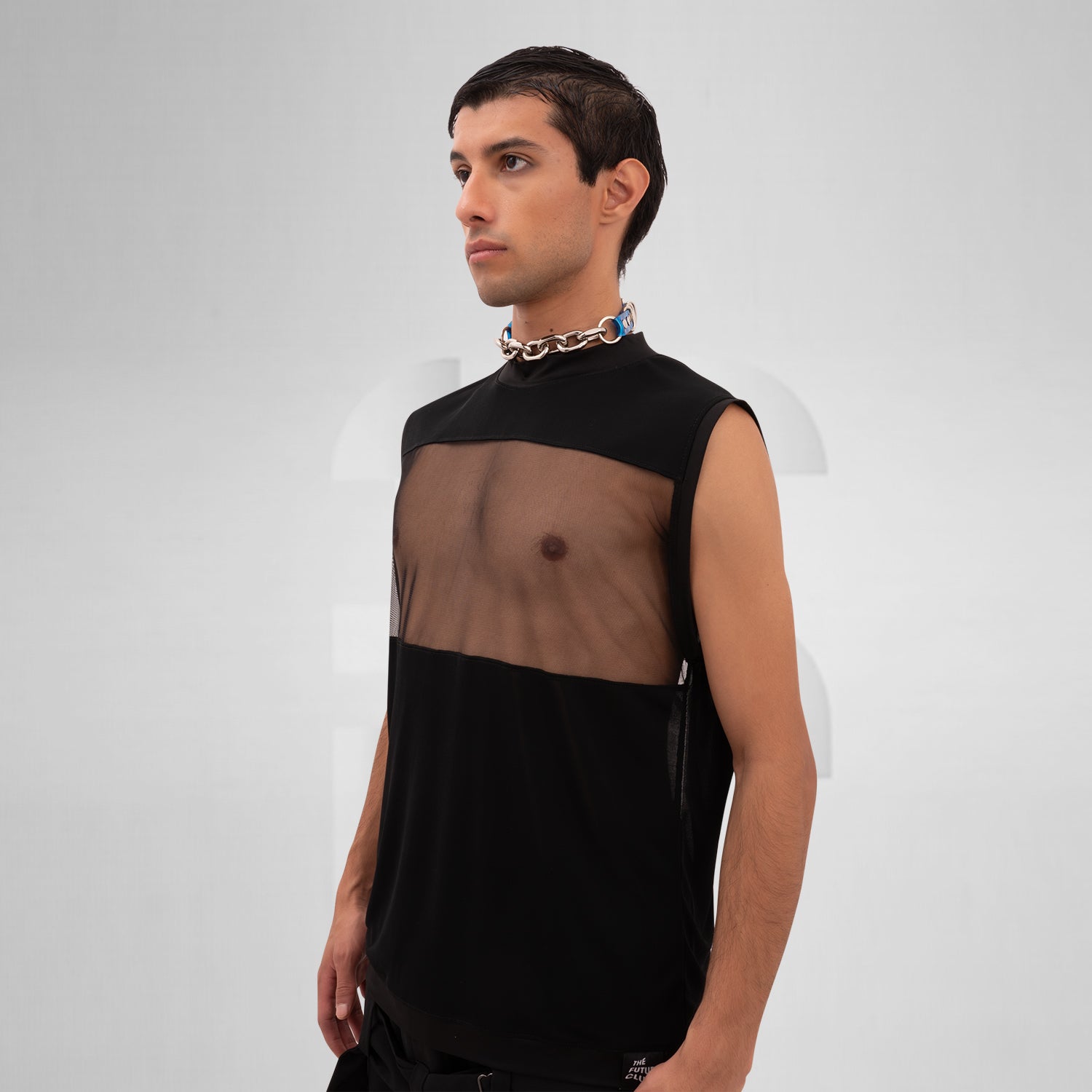 A male model is wearing a black sleeveless tank top with a bold mesh panel across the chest, revealing part of the upper torso. The tank top has a high neckline and a minimalist, modern design, paired with a chunky silver chain necklace. The background is plain and minimal, focusing attention on the garment's details.