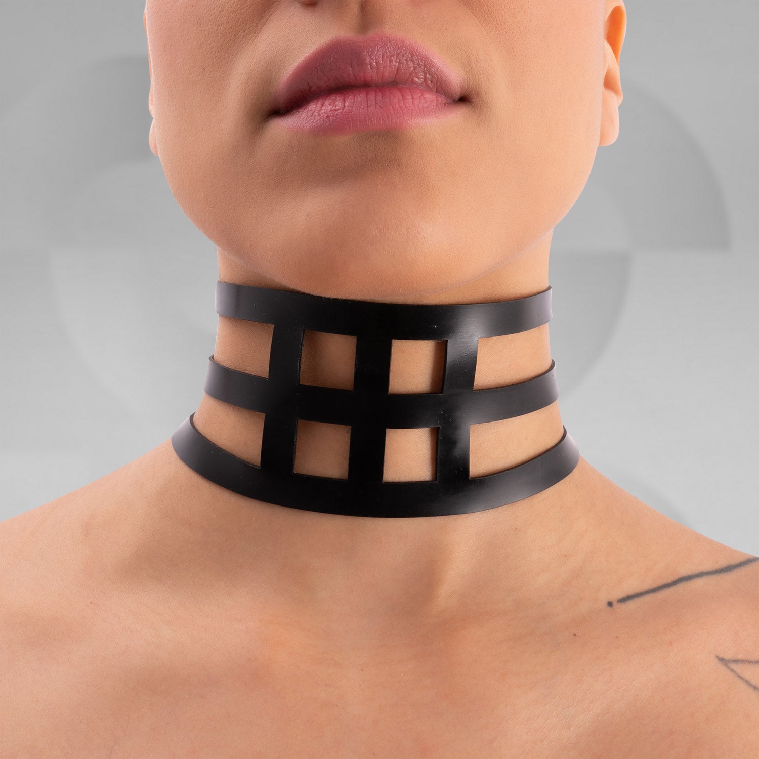 The image features a close-up of a person wearing a bold black latex choker with an intricate grid design. The statement piece elegantly wraps around the neck, adding a striking visual element to the overall look. The model's minimalistic makeup and shaved head emphasize the choker's edgy aesthetic, making it a perfect accessory for both avant-garde fashion and daring street style. This unique design showcases individuality and confidence, ideal for making a memorable impression.