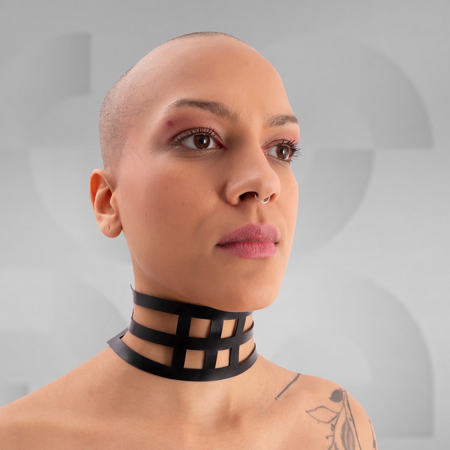 The image features a close-up of a person wearing a bold black latex choker with an intricate grid design. The statement piece elegantly wraps around the neck, adding a striking visual element to the overall look. The model's minimalistic makeup and shaved head emphasize the choker's edgy aesthetic, making it a perfect accessory for both avant-garde fashion and daring street style. This unique design showcases individuality and confidence, ideal for making a memorable impression.