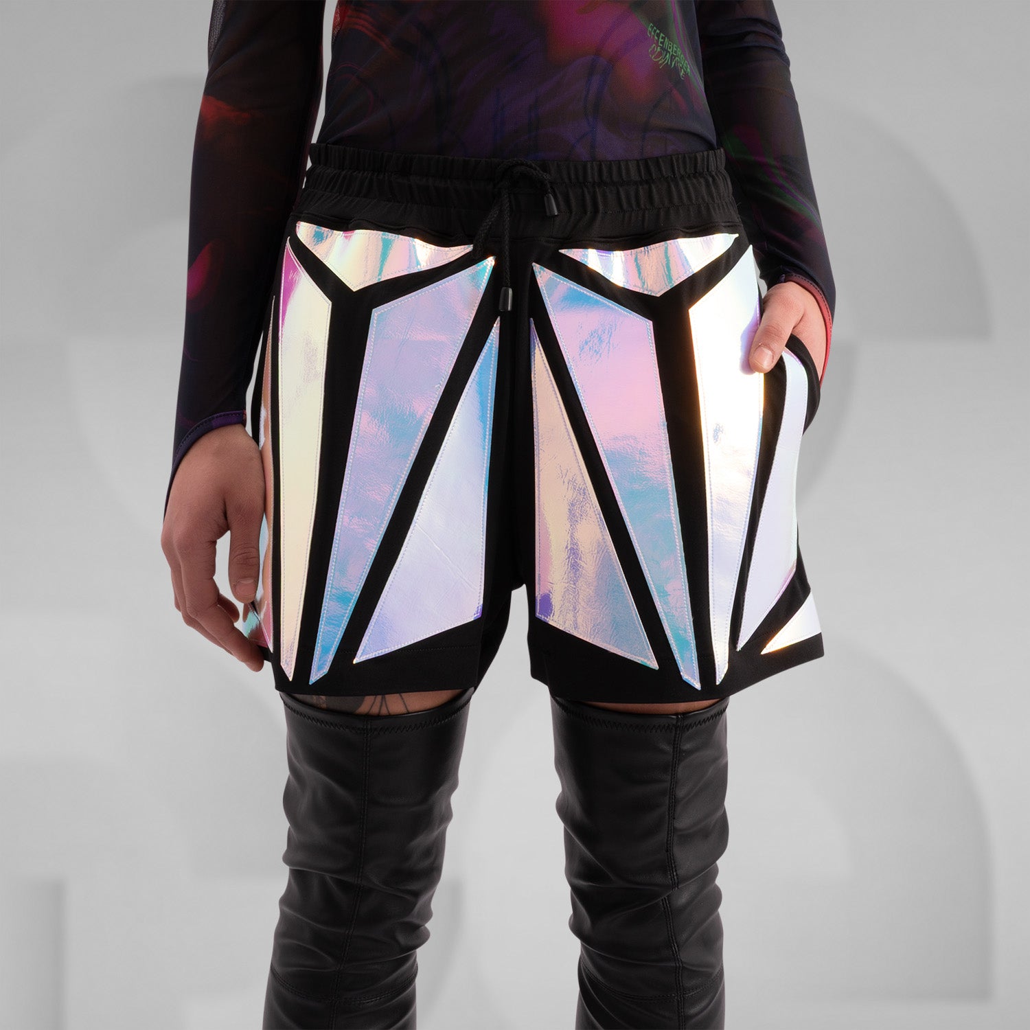 Close-up of a model wearing black shorts with striking holographic geometric panels, reflecting iridescent colors. Paired with a sheer long-sleeve top and thigh-high black leather boots, this futuristic and edgy look is perfect for festival, rave, and avant-garde fashion.