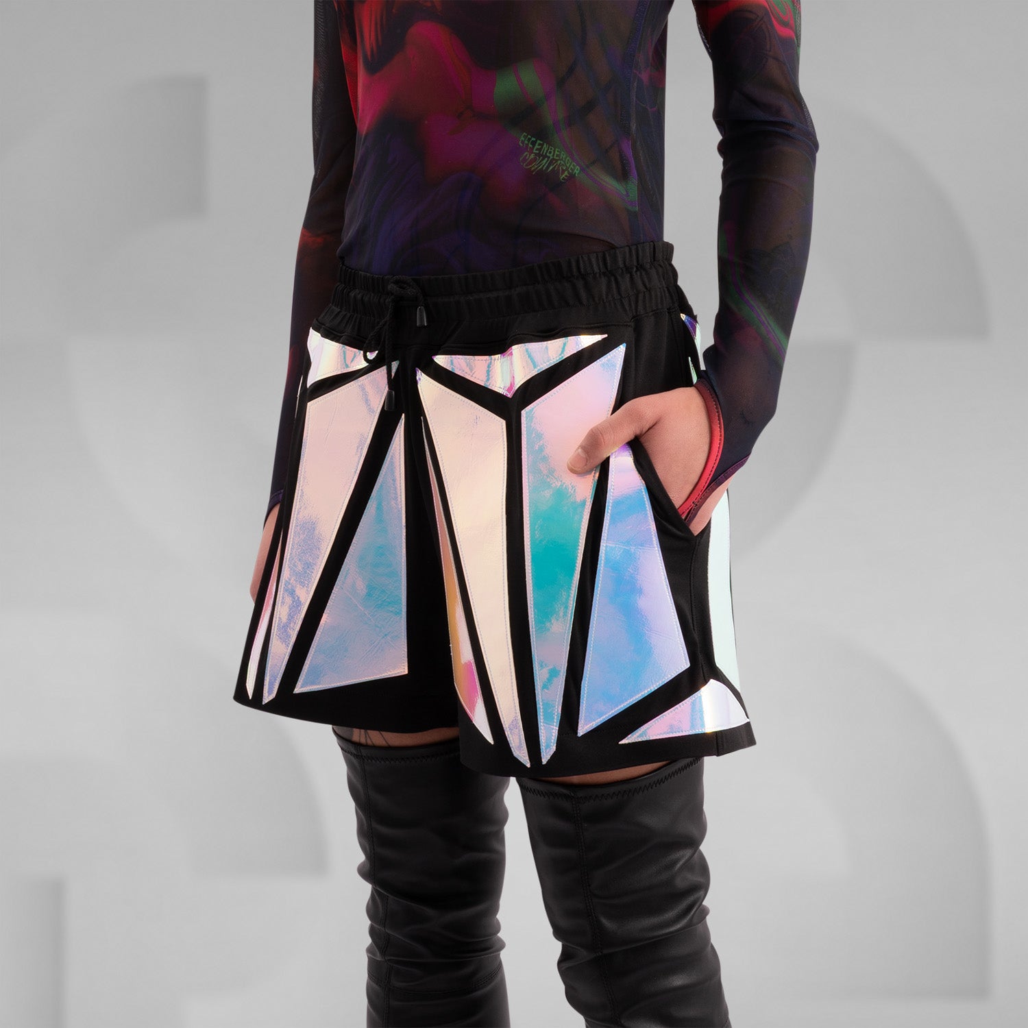Close-up of a model wearing black shorts with striking holographic geometric panels, reflecting iridescent colors. Paired with a sheer long-sleeve top and thigh-high black leather boots, this futuristic and edgy look is perfect for festival, rave, and avant-garde fashion.
