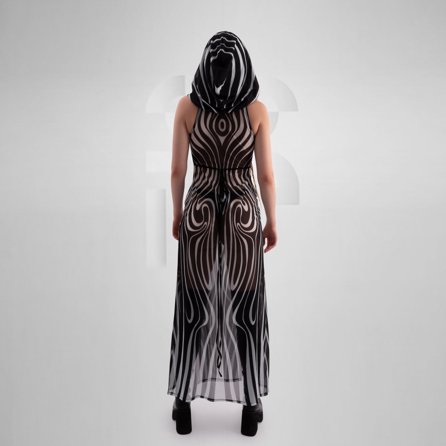 Model wearing a sleeveless, sheer black and white optical illusion print mesh dress with a high neck and cut-out chest detail, styled with black combat boots, standing confidently against a minimalist grey background everything looks very tribal festival like.