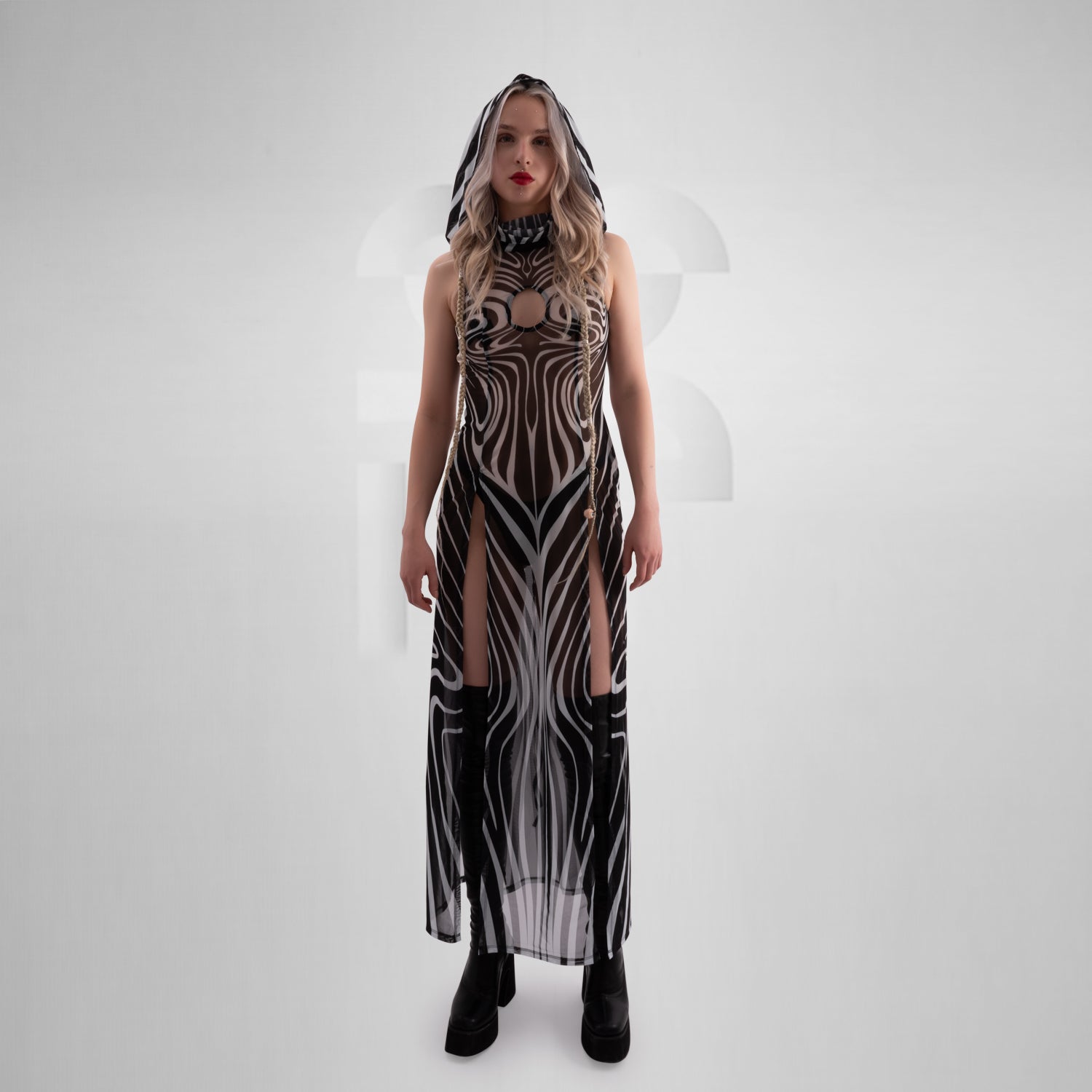 Model wearing a sleeveless, sheer black and white optical illusion print mesh dress with a high neck and cut-out chest detail, styled with black combat boots, standing confidently against a minimalist grey background everything looks very tribal festival like.