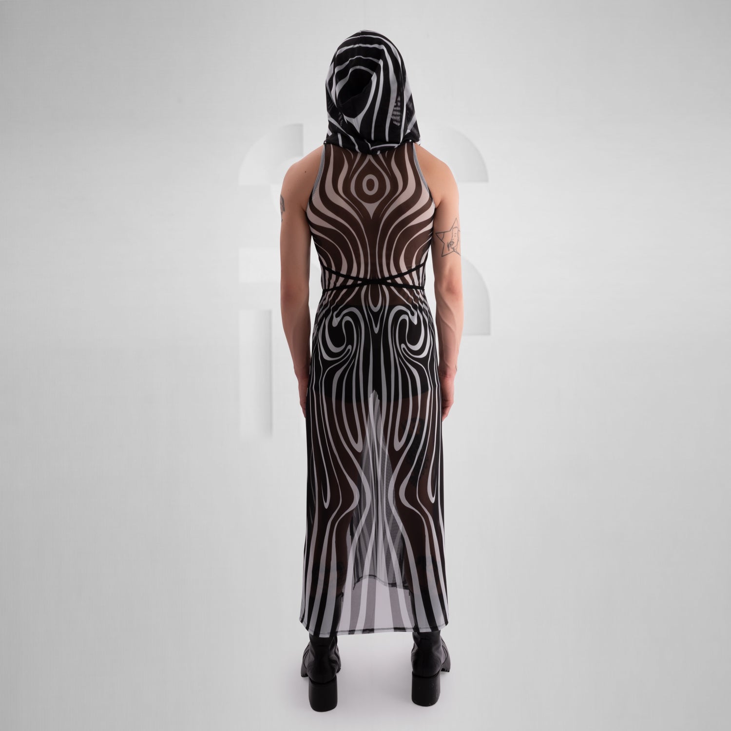 Model wearing a sleeveless, sheer black and white optical illusion print mesh dress with a high neck and cut-out chest detail, styled with black combat boots, standing confidently against a minimalist grey background everything looks very tribal festival like.
