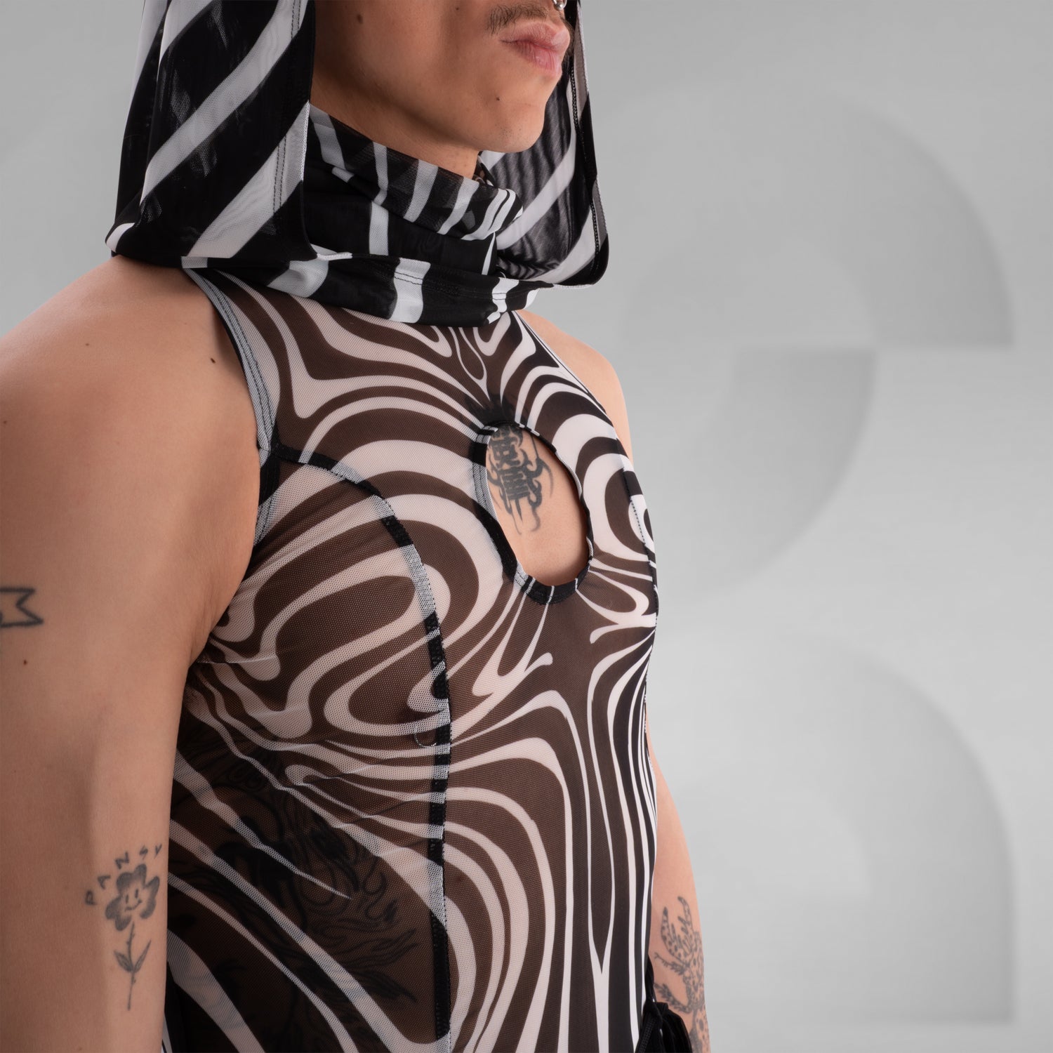 Model wearing a sleeveless, sheer black and white optical illusion print mesh dress with a high neck and cut-out chest detail, styled with black combat boots, standing confidently against a minimalist grey background everything looks very tribal festival like.