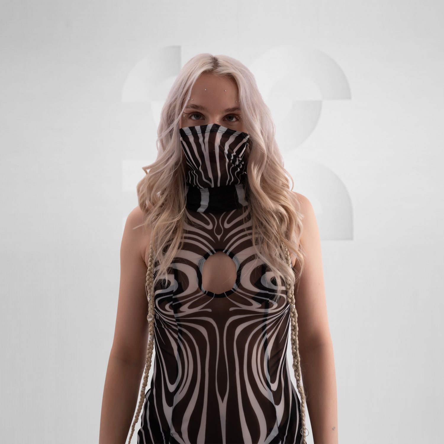 Model wearing a sleeveless, sheer black and white optical illusion print mesh dress with a high neck and cut-out chest detail, styled with black combat boots, standing confidently against a minimalist grey background everything looks very tribal festival like.