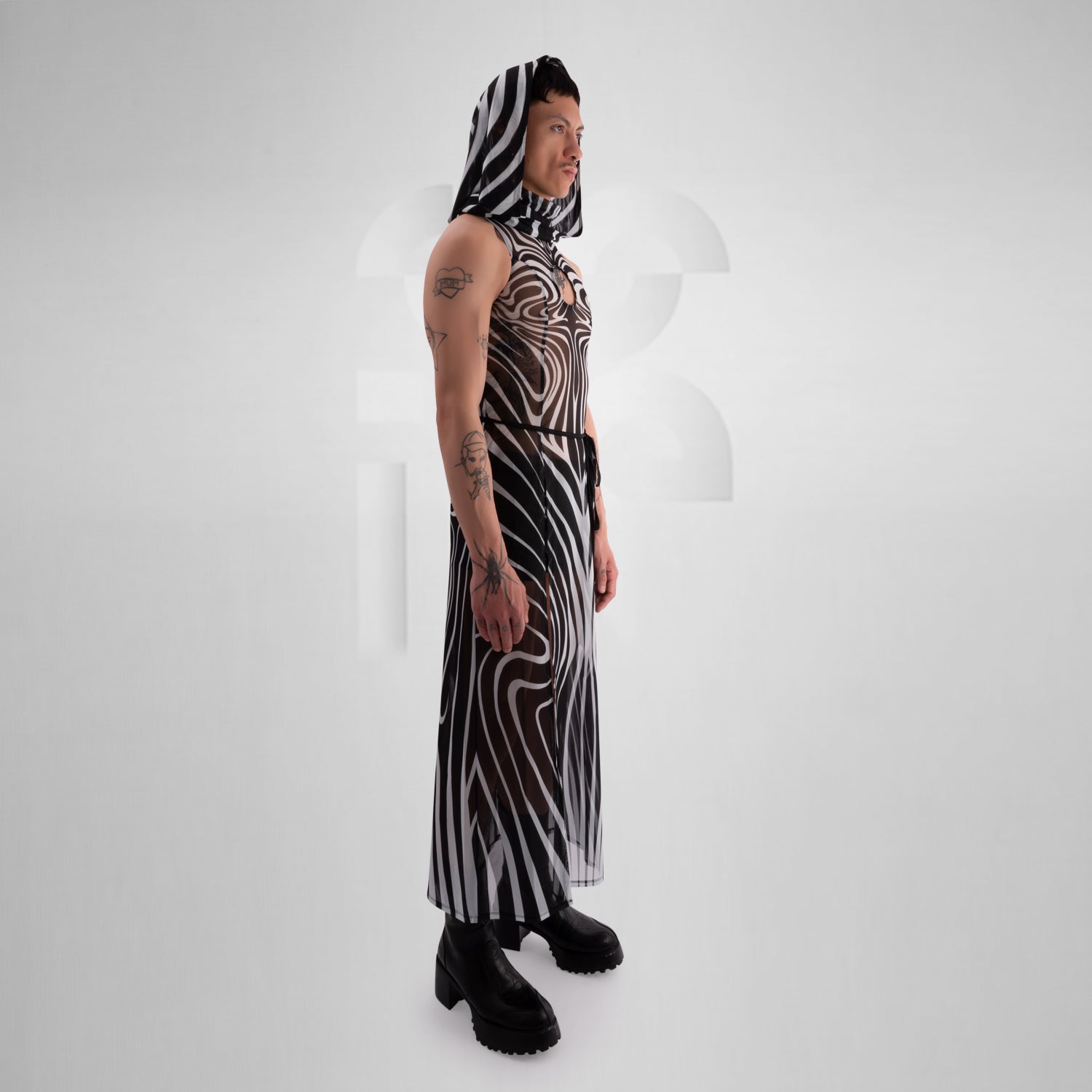 Model wearing a sleeveless, sheer black and white optical illusion print mesh dress with a high neck and cut-out chest detail, styled with black combat boots, standing confidently against a minimalist grey background everything looks very tribal festival like.