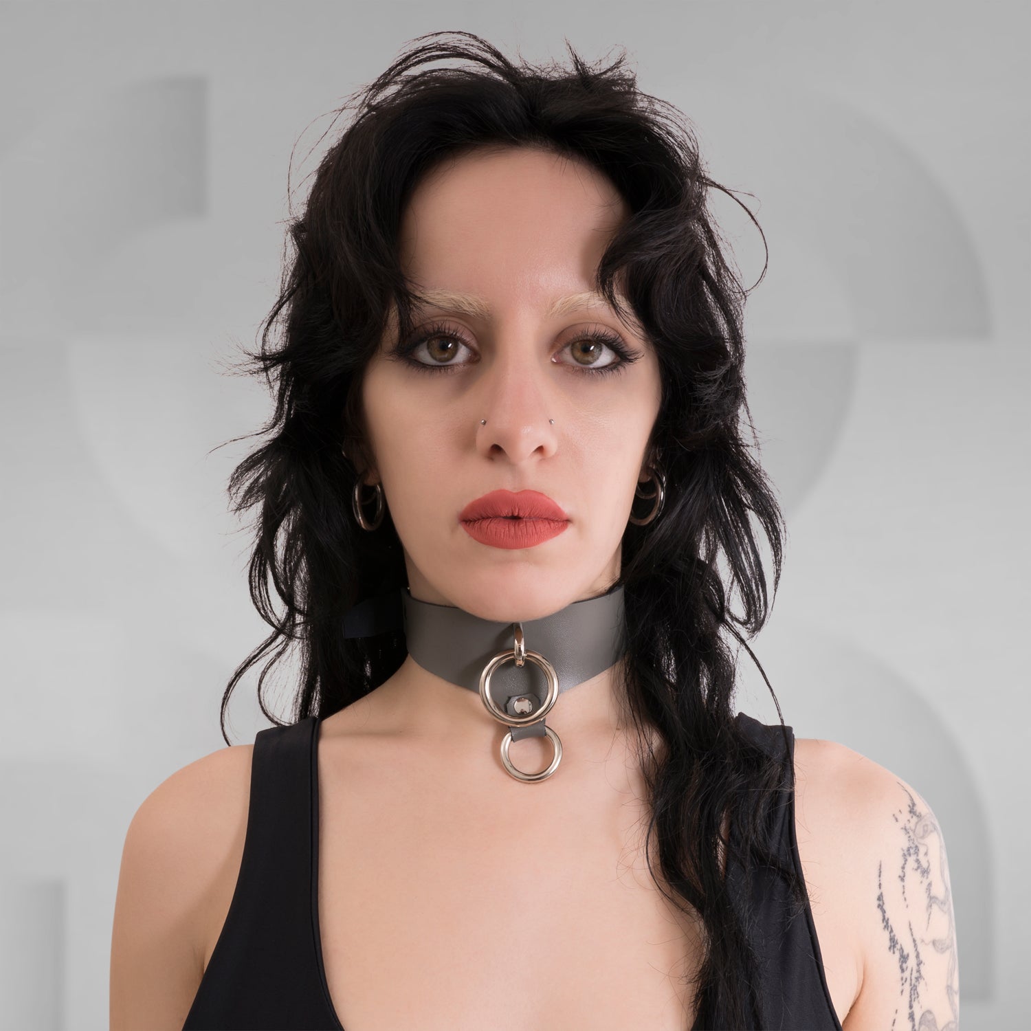 a sleek gray choker featuring circular metallic details that add an edgy touch to her look. The minimalist design is complemented by her dark hair and makeup, creating a striking, modern aesthetic. The neutral background emphasizes her bold features and the unique accessories.